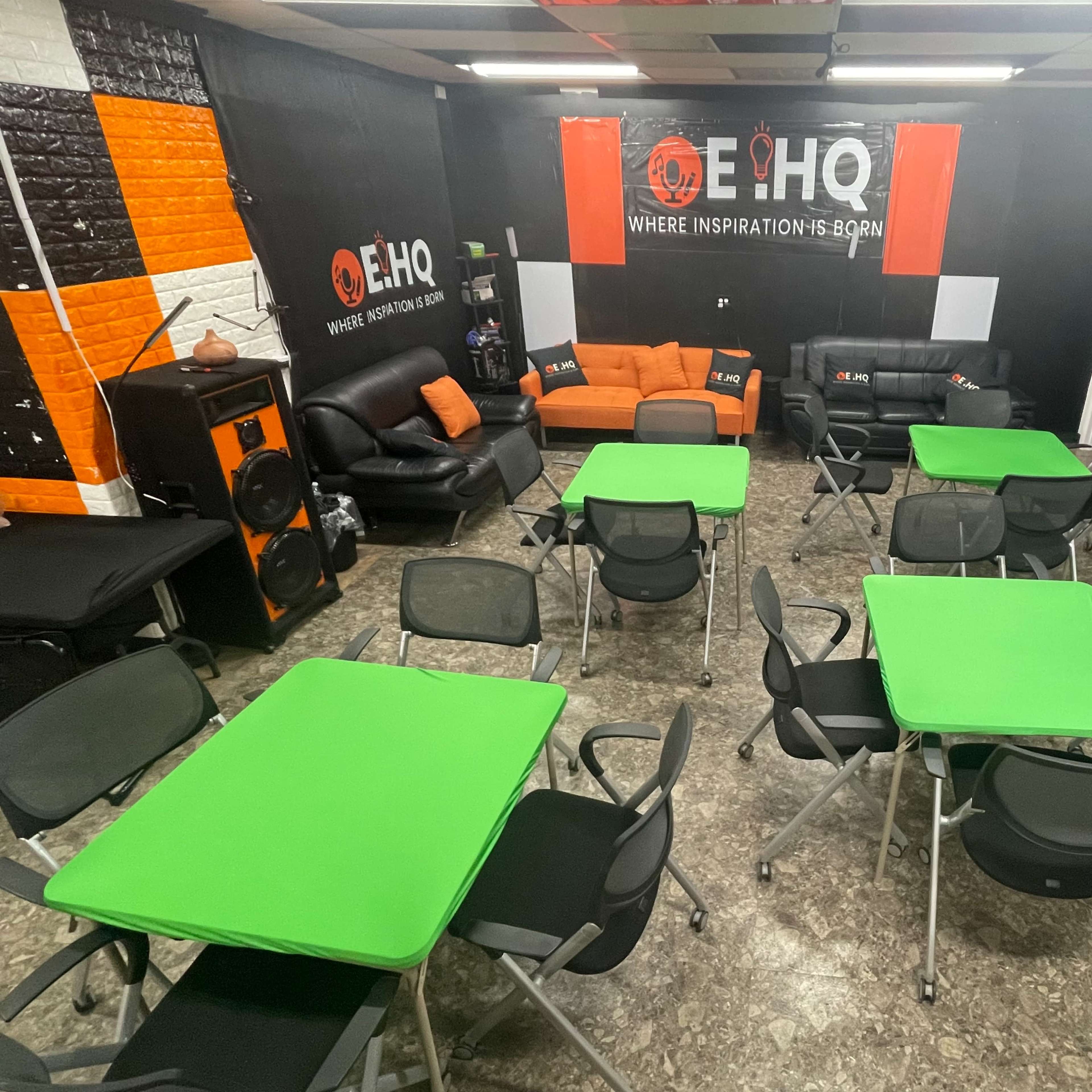 A spacious room arranged with several green tables and black chairs, featuring a lounge area with orange and black seating against a branded wall.