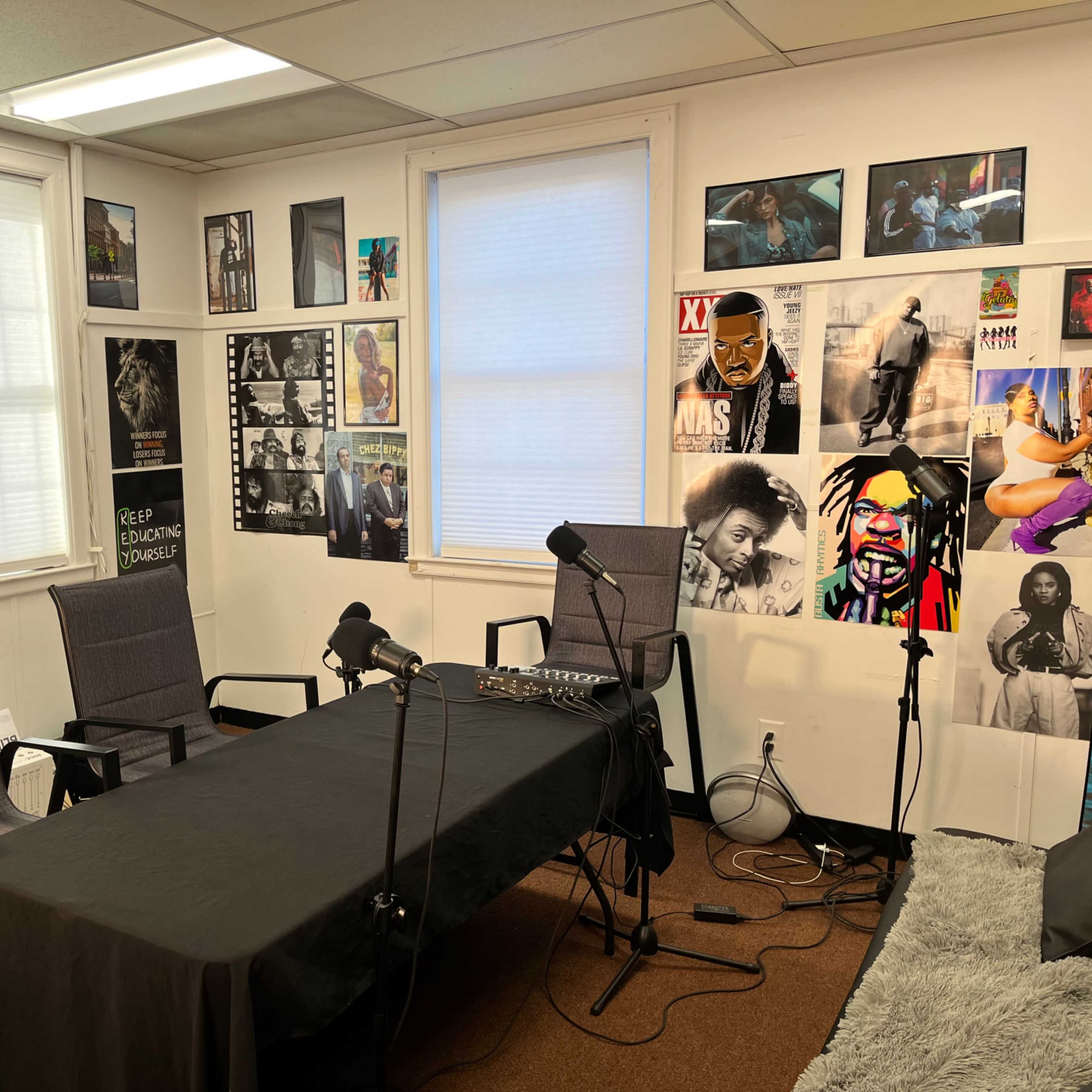 A small room set up for a recording or podcast, featuring a black table, three chairs, microphones, and various photographs on the walls.