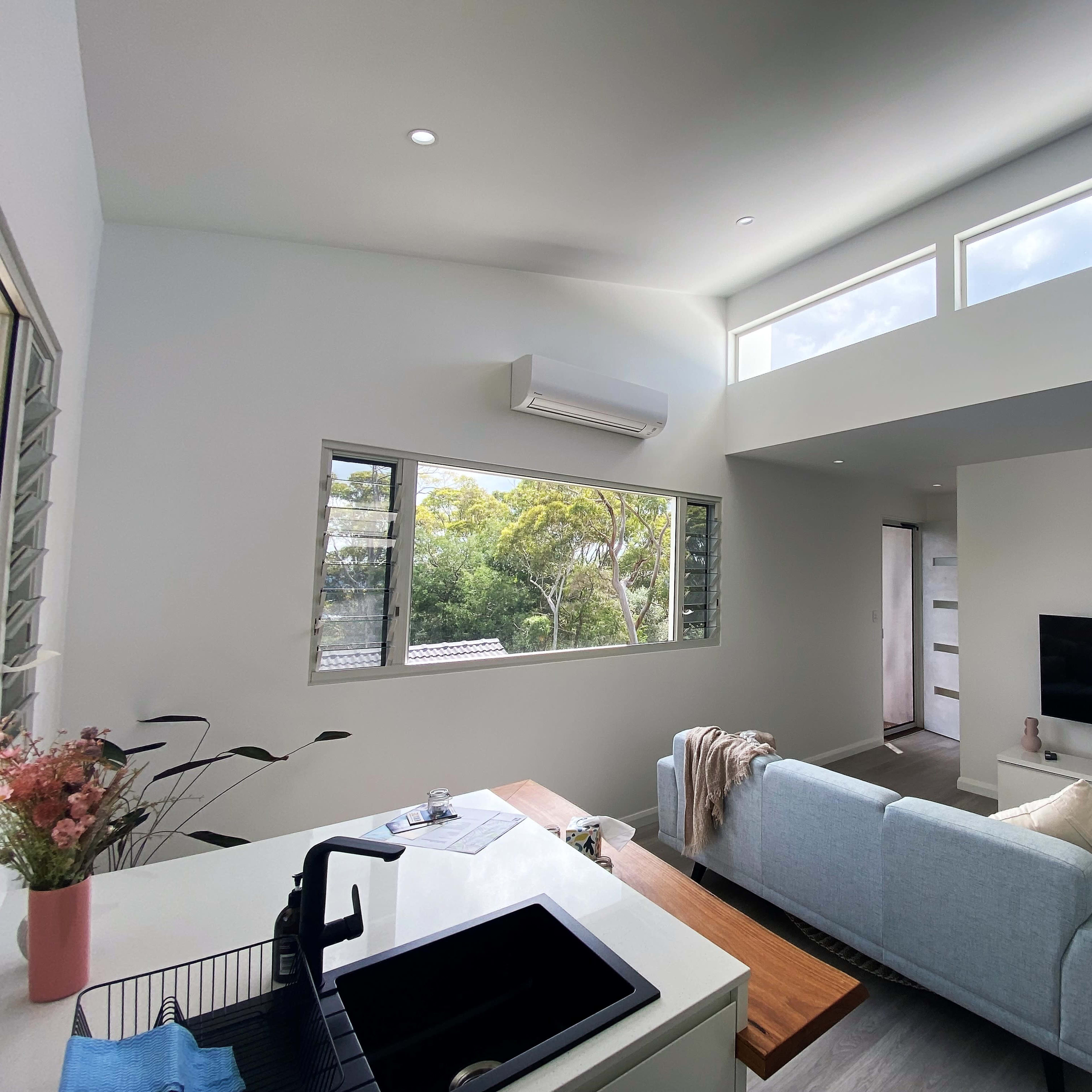 A modern living room with large windows, a light-colored sofa, a television mounted on the wall, and a kitchen area featuring a sink and countertop.