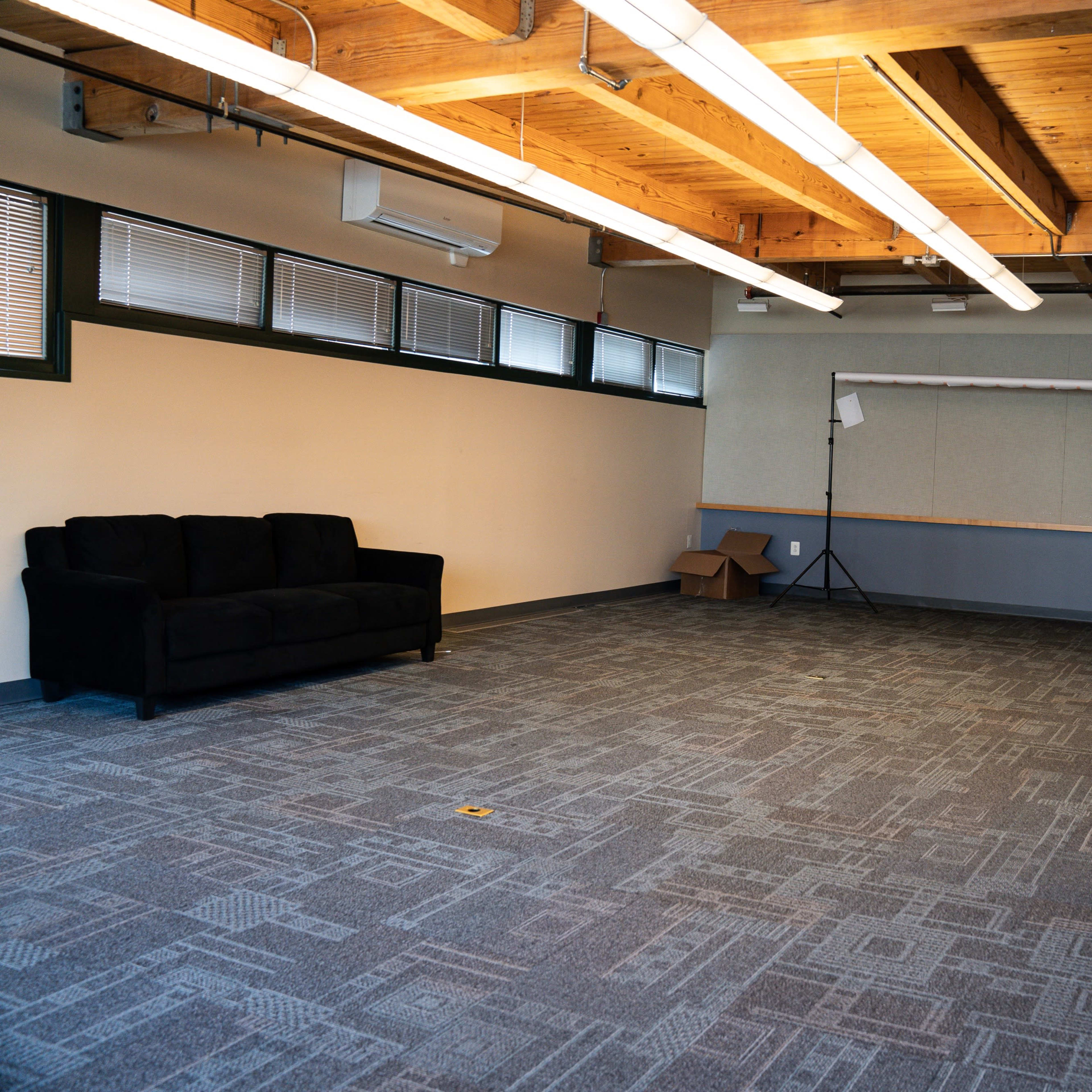 An empty room with wooden beams, a black couch, a folding backdrop setup, and a desk with a stool.