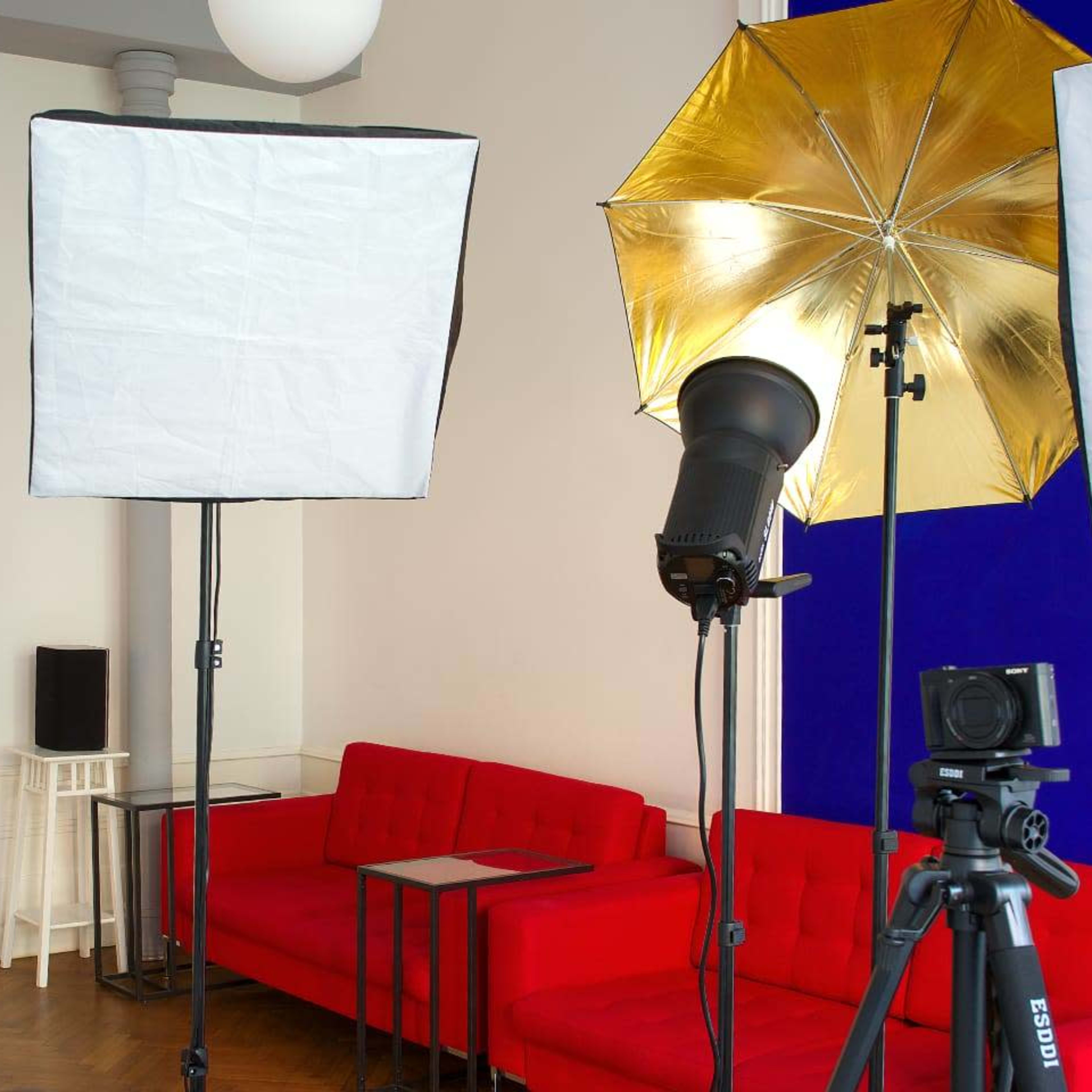 A bright studio setup featuring three photography lighting rigs and two cameras facing a red sofa and a small side table.