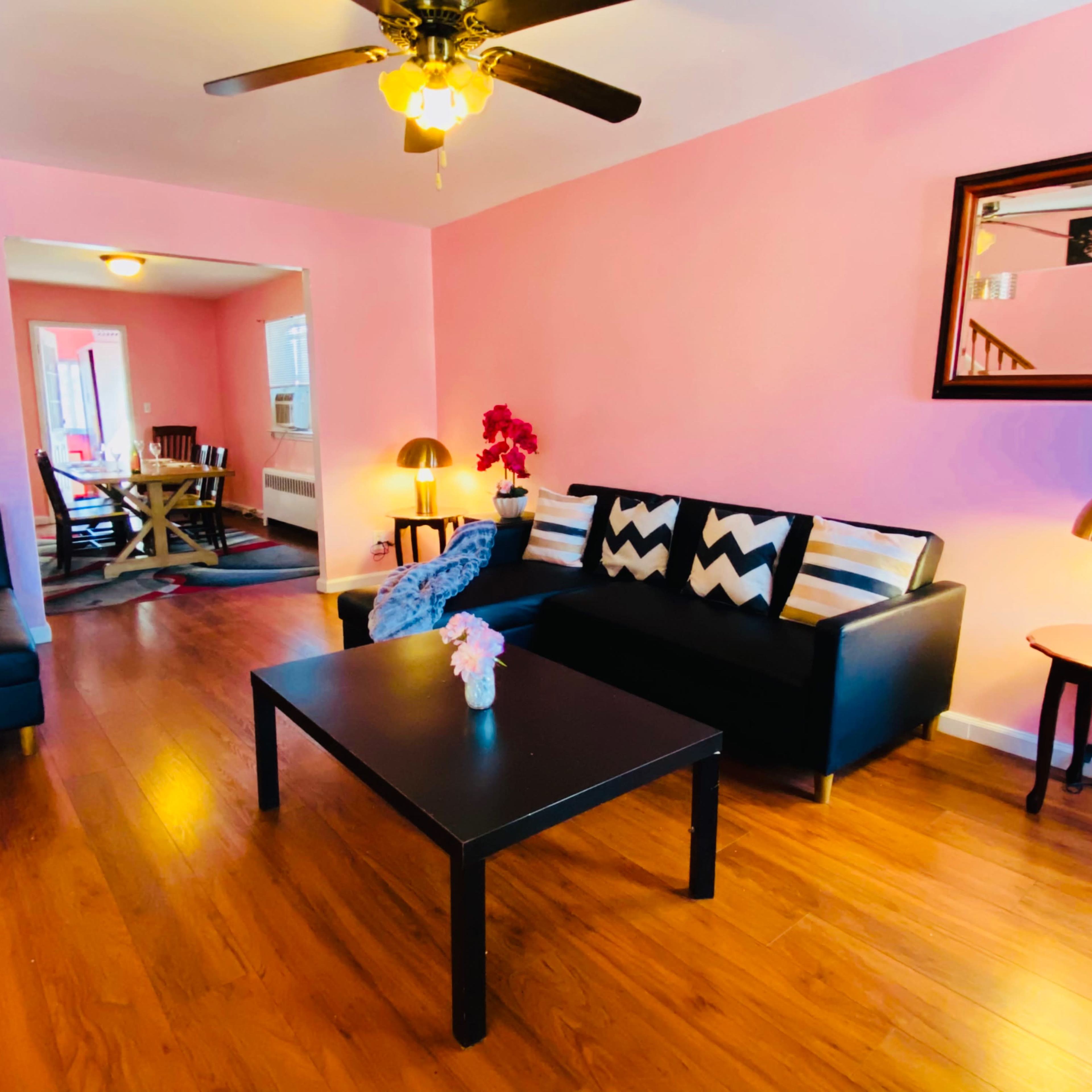 A living room with pink walls, a black sofa and coffee table, and a dining area visible in the background.