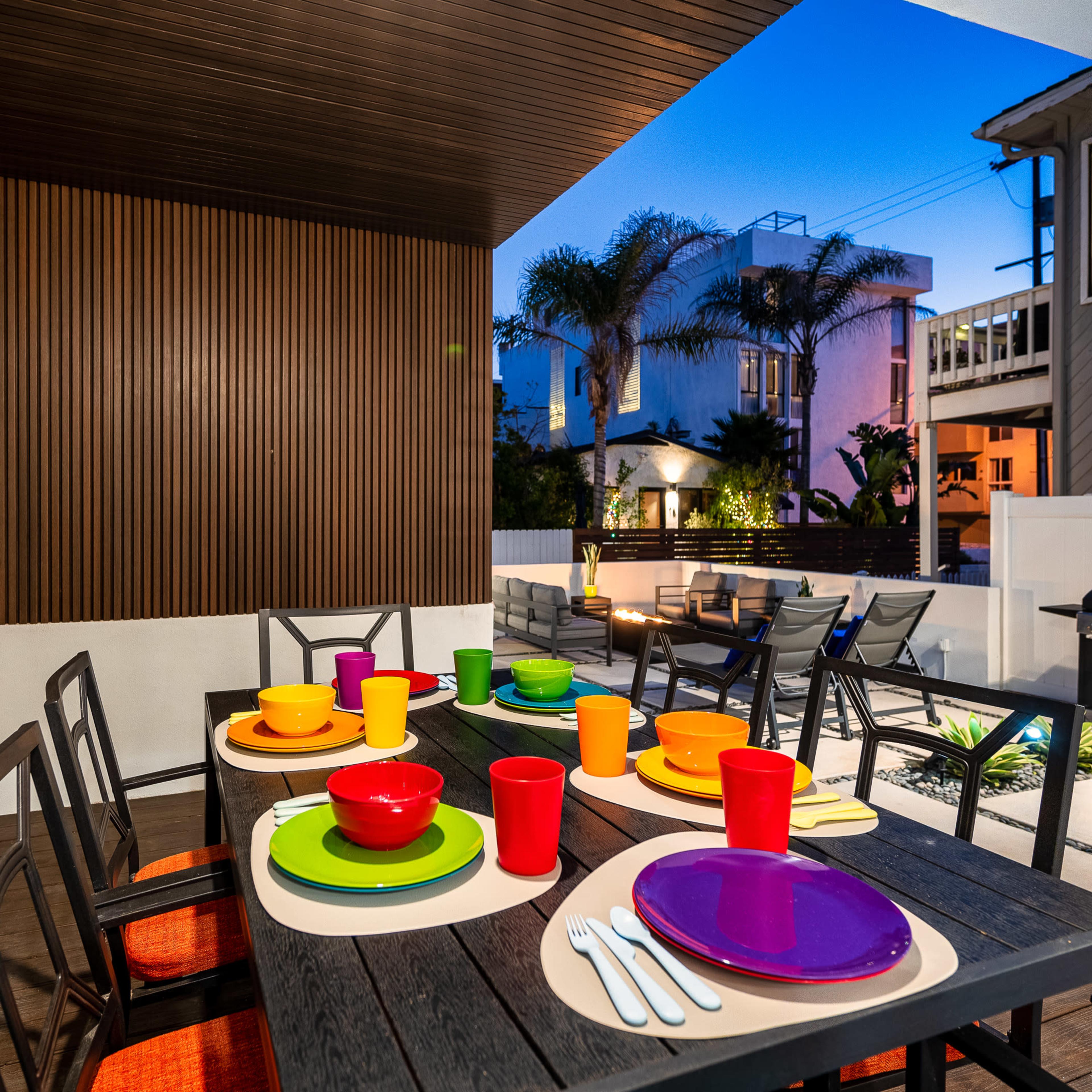A brightly colored outdoor dining area features a wooden table set with plates and cups, surrounded by modern chairs and a chimney in the background.