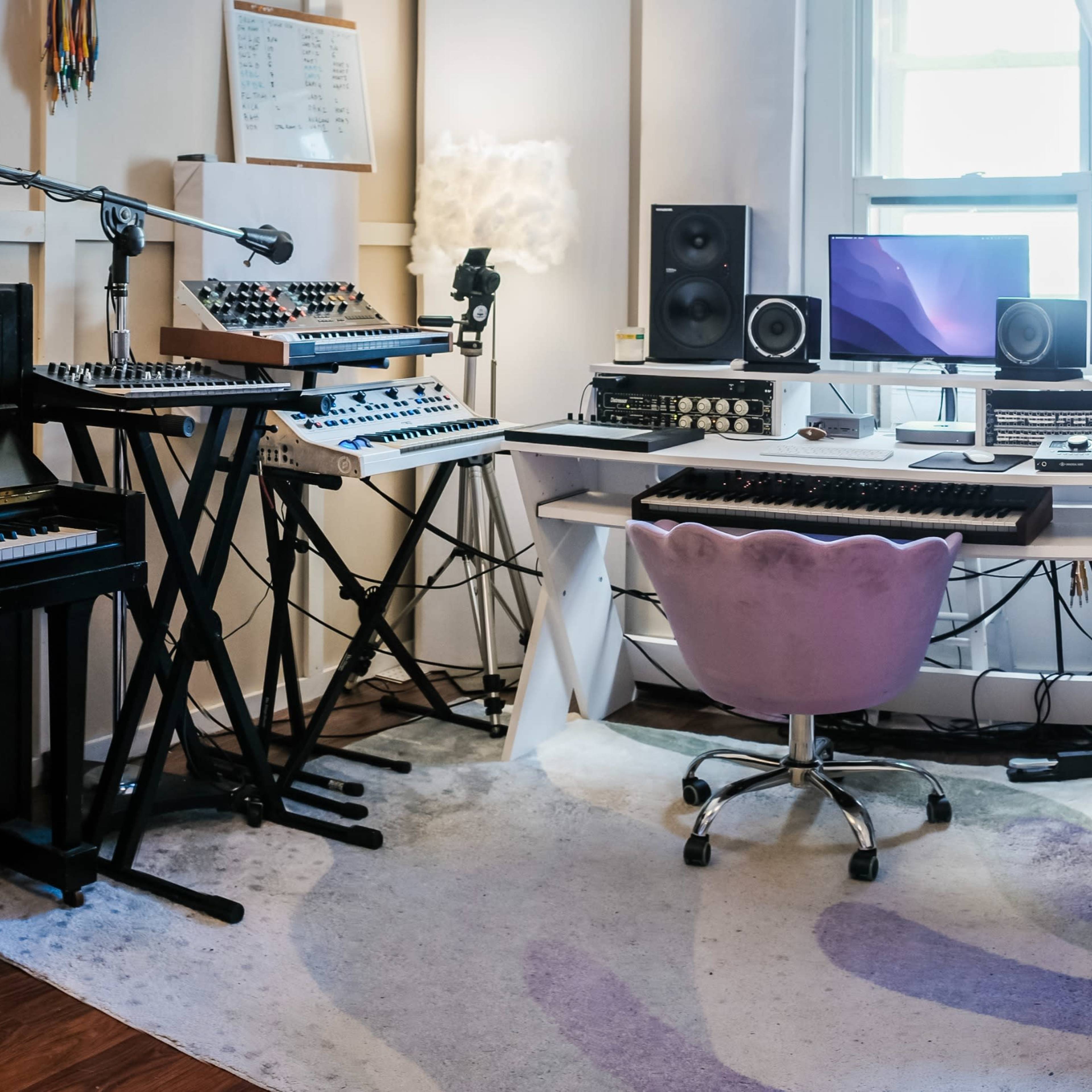 A music studio with a black piano, synthesizer, computer, monitors, and various equipment on a desk.