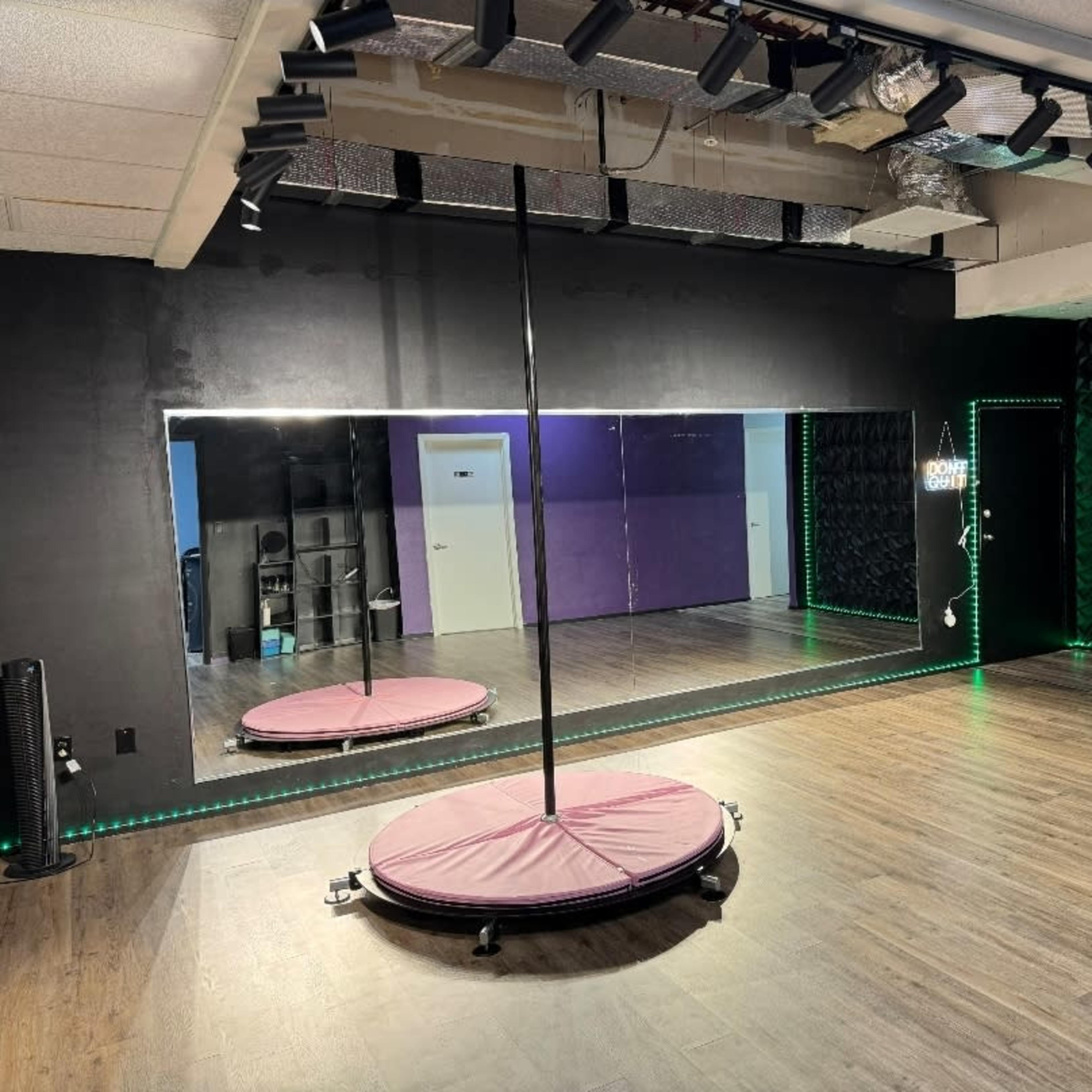 A dance studio featuring a mirrored wall, a central pole, and LED lighting along the edges of the room.