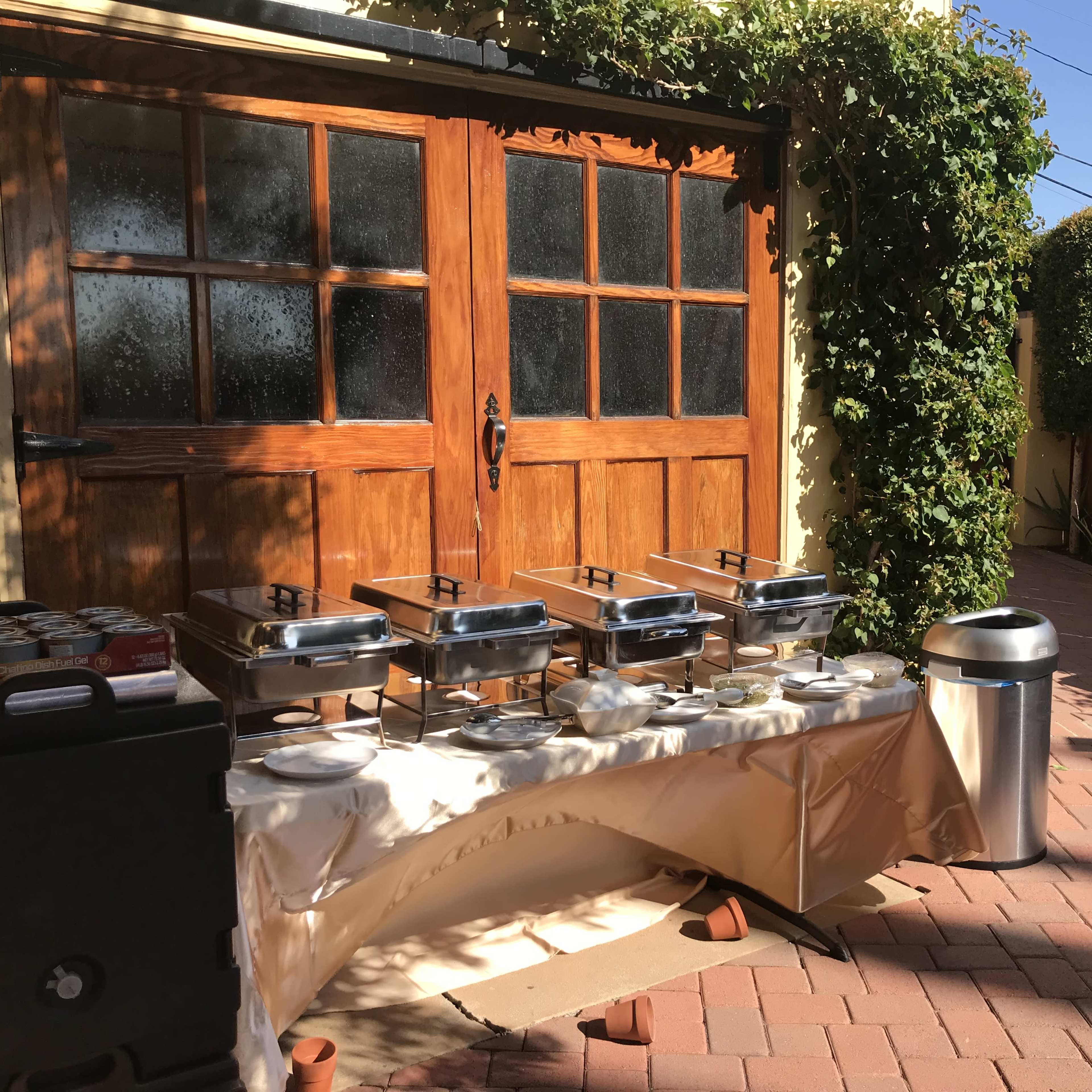 A buffet setup with metal chafing dishes, plates, and a trash can is arranged in front of wooden doors in a sunny outdoor area.