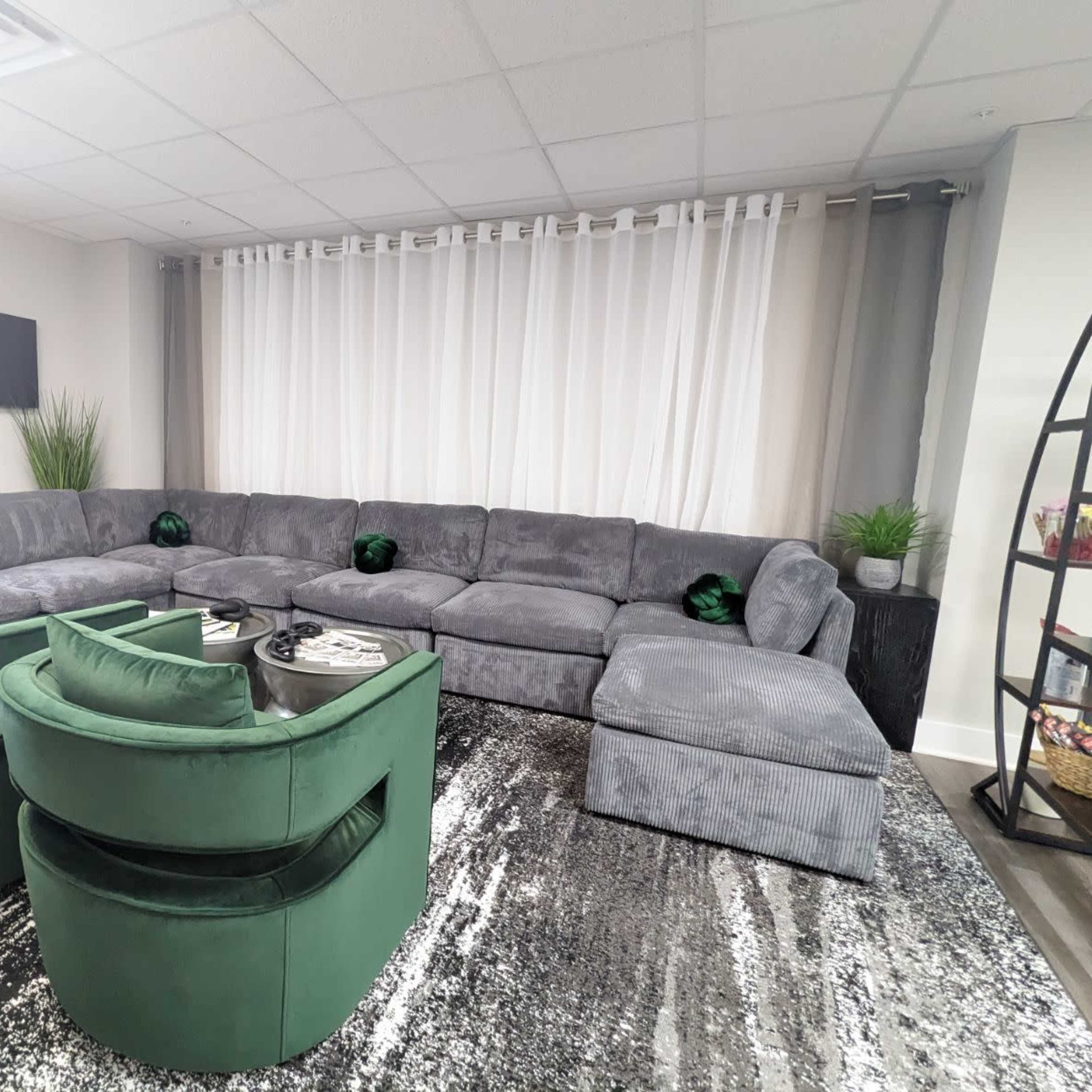A modern lounge area featuring a large gray sectional sofa, two green velvet chairs, and a black and white patterned rug.