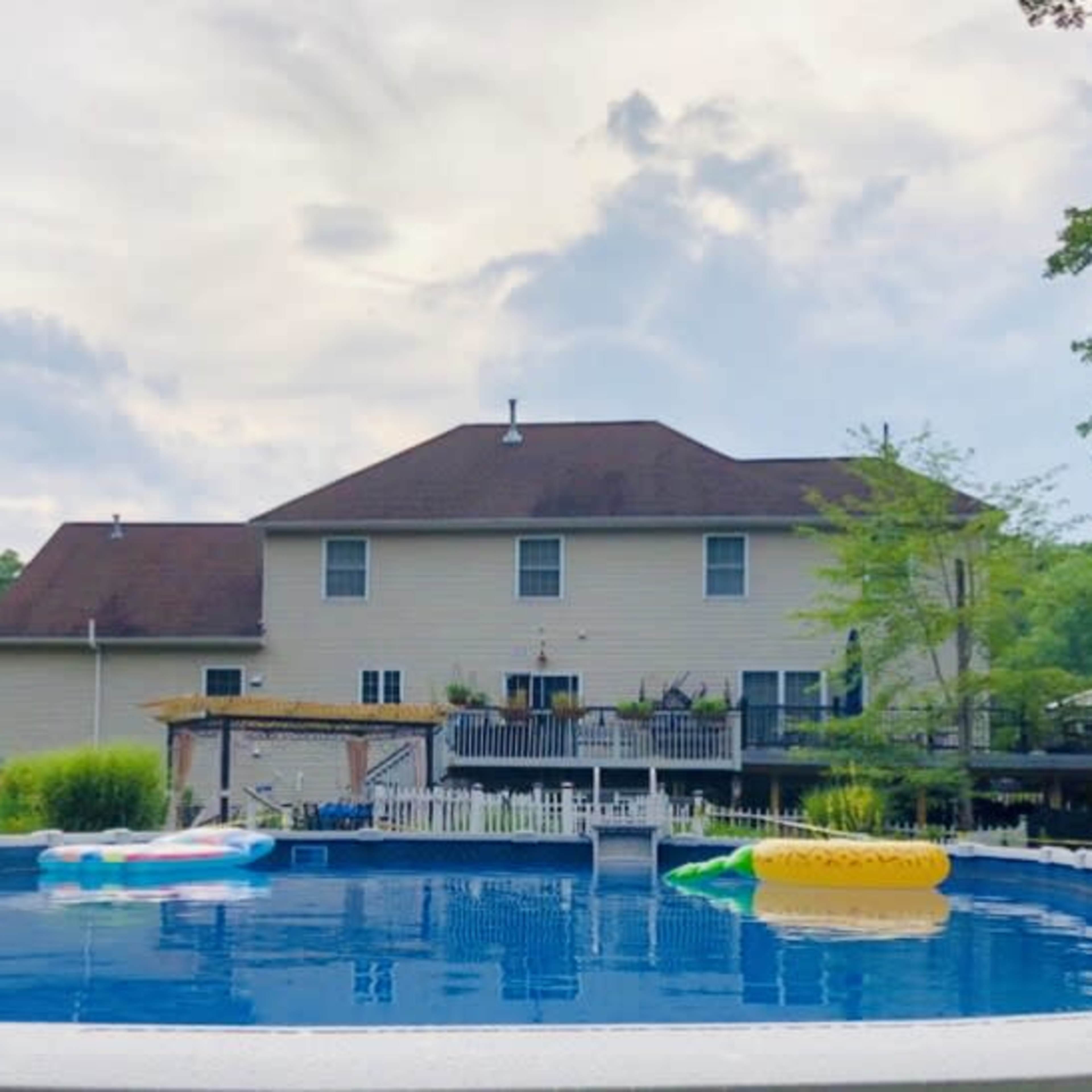 A large above-ground pool is surrounded by trees and vegetation, with a two-story house featuring a deck visible in the background.