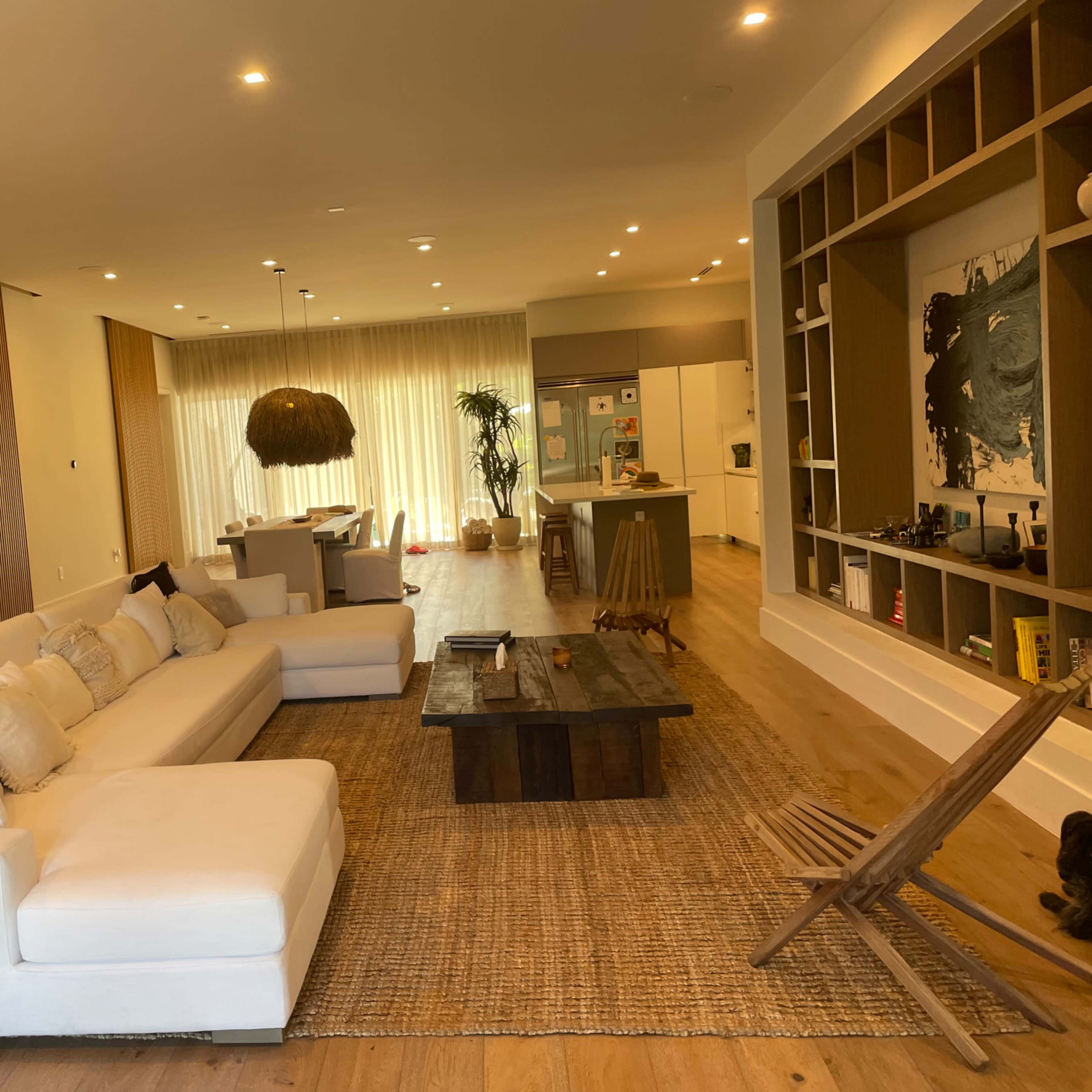 A modern living room with a large white sectional sofa, a wooden coffee table, and a woven rug, leading into a kitchen area with multiple plants and ample natural light.