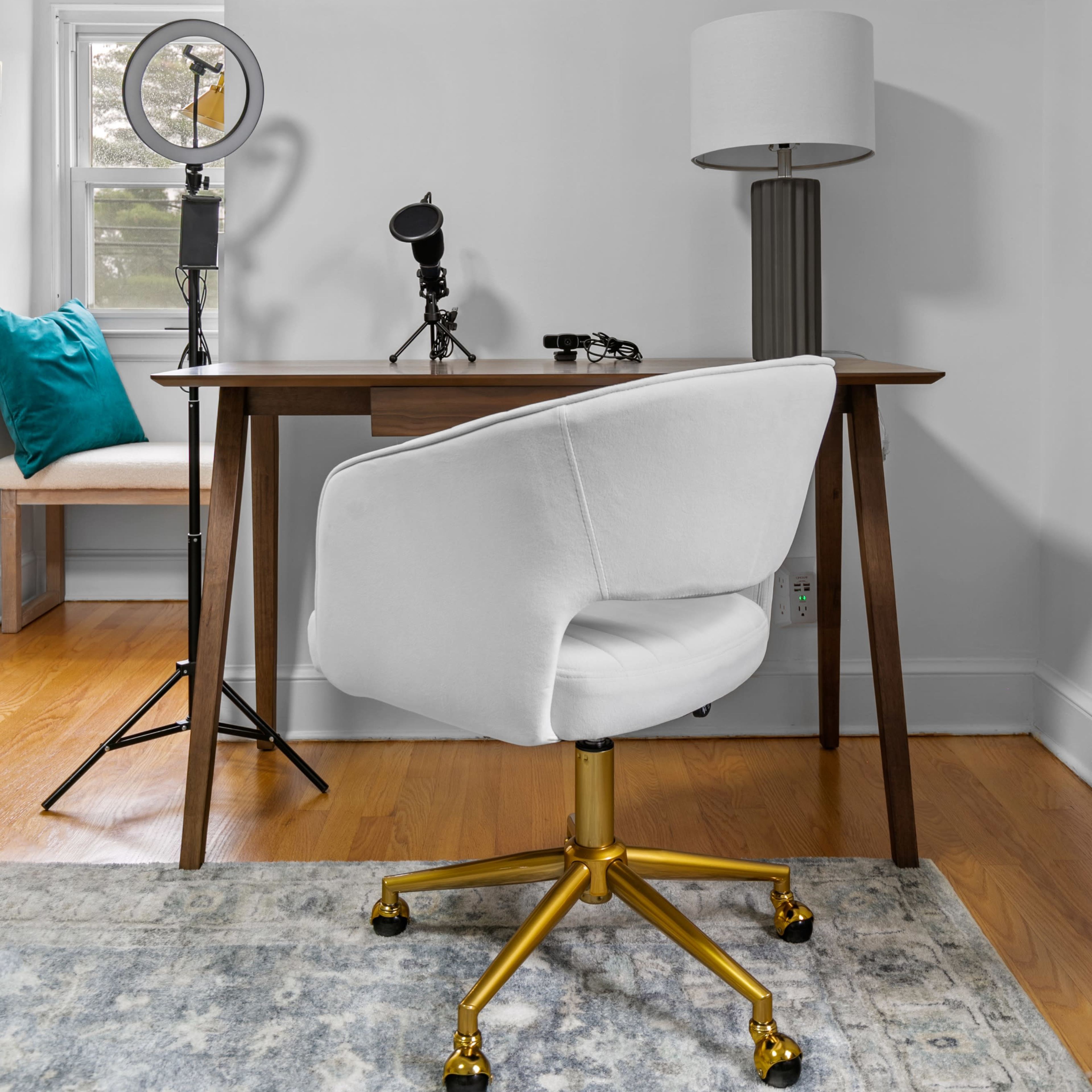 A modern home office features a wooden desk, a white swivel chair with a gold base, a lamp, and a ring light setup beside it.
