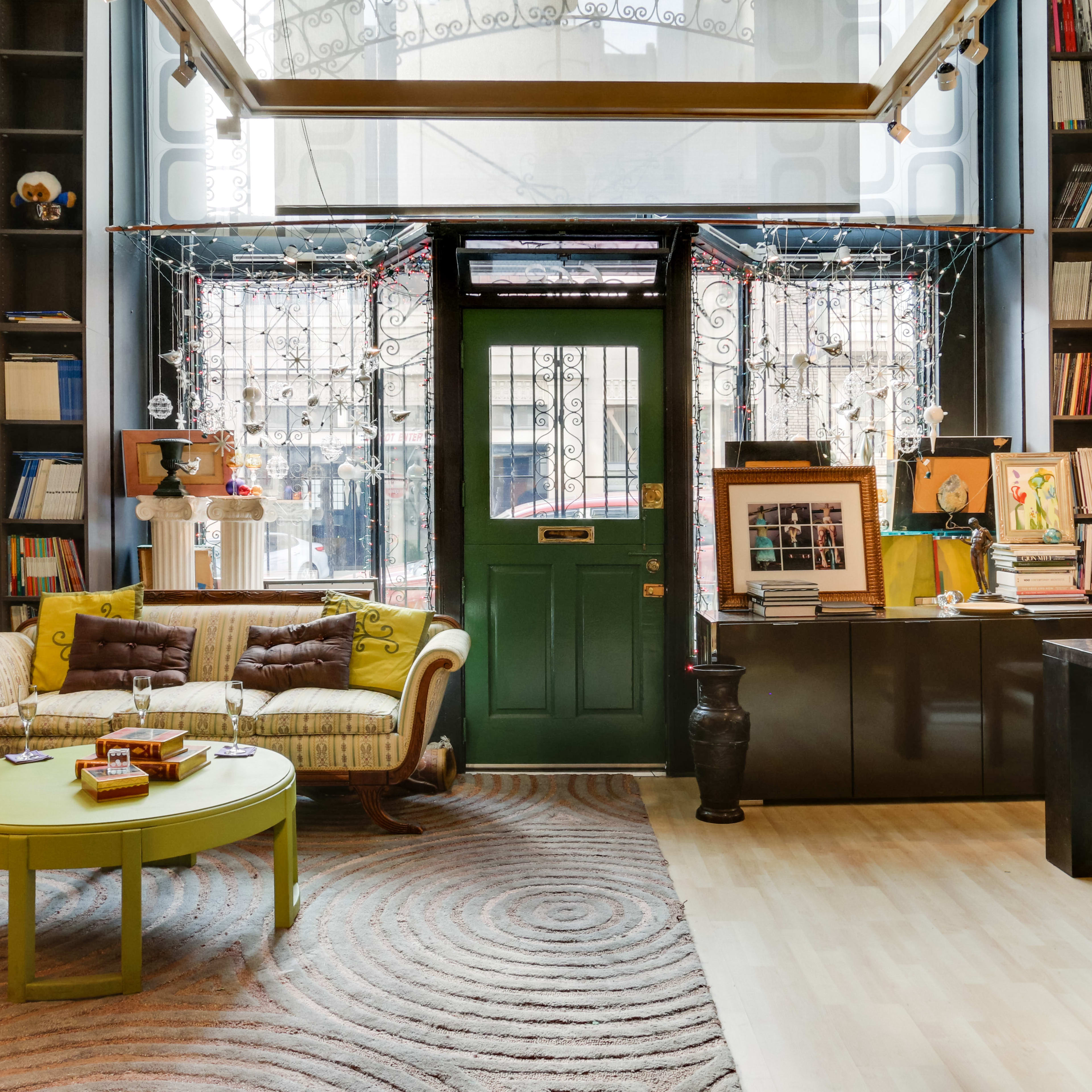 A cozy interior of a room featuring a green door, a yellowish sofa with a circular coffee table, and walls adorned with framed pictures and shelves filled with books and art.