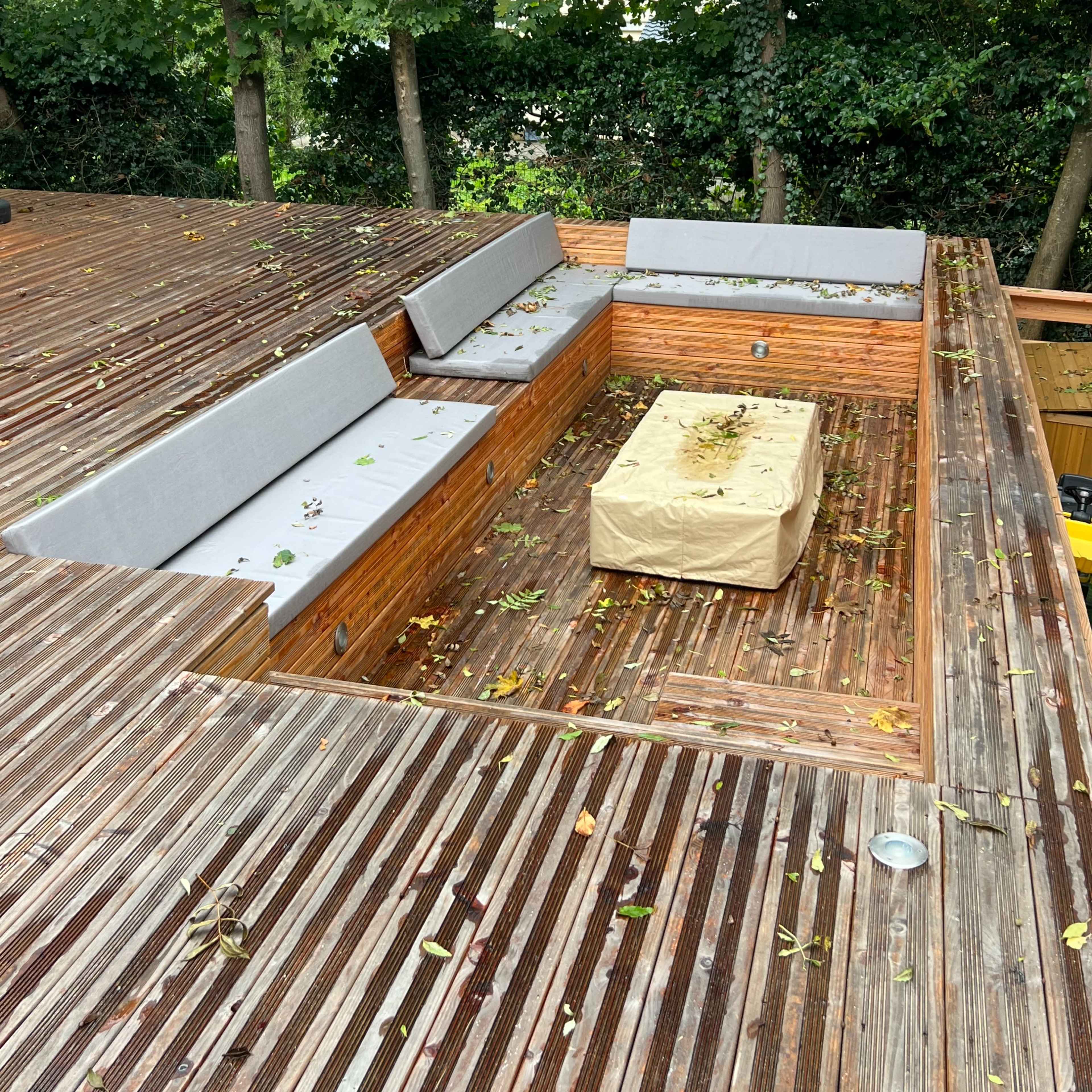 A wooden terrace features a sunken seating area with gray cushions and a covered table, surrounded by trees.