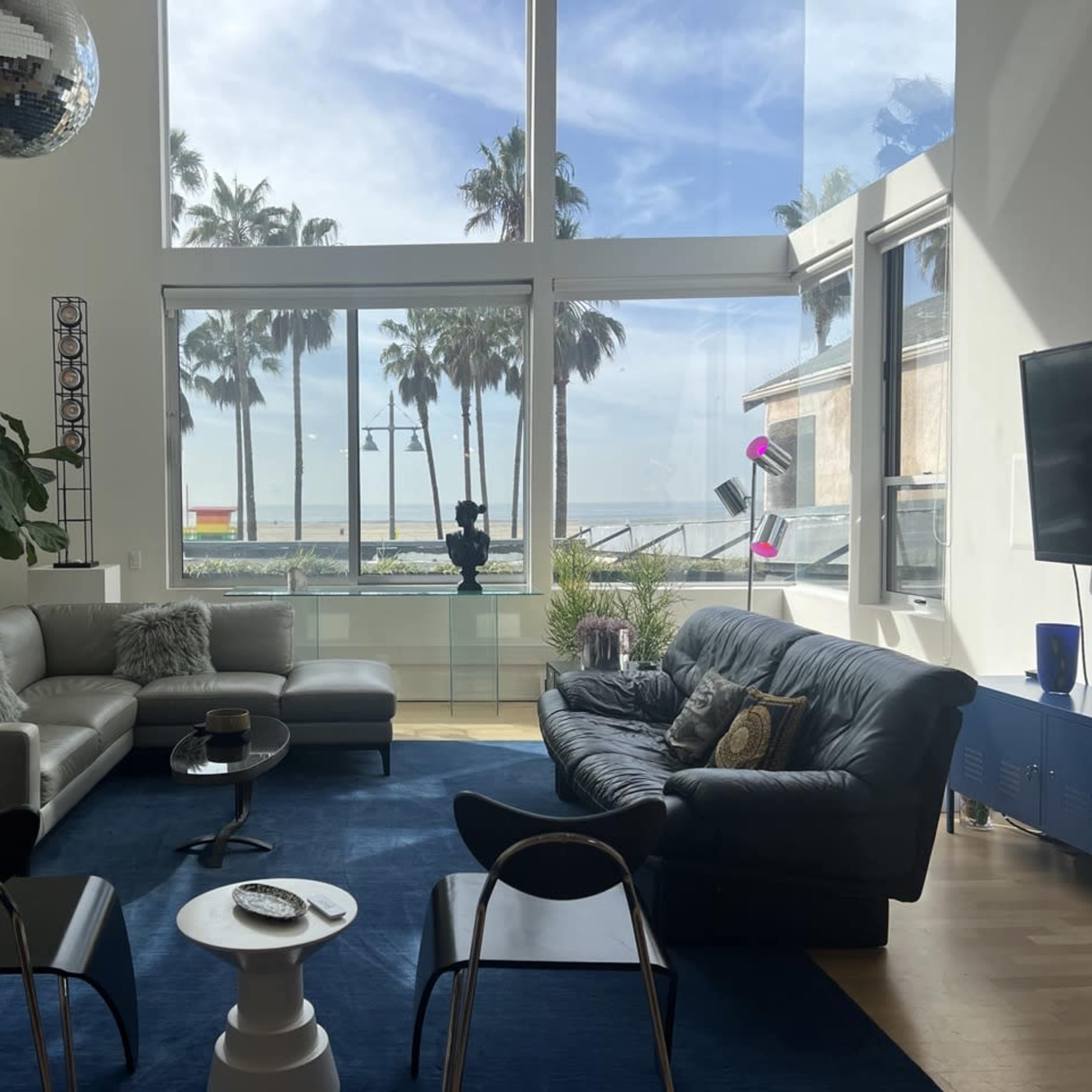 A modern living room features two sofas, a coffee table, and large windows overlooking a beach with palm trees.