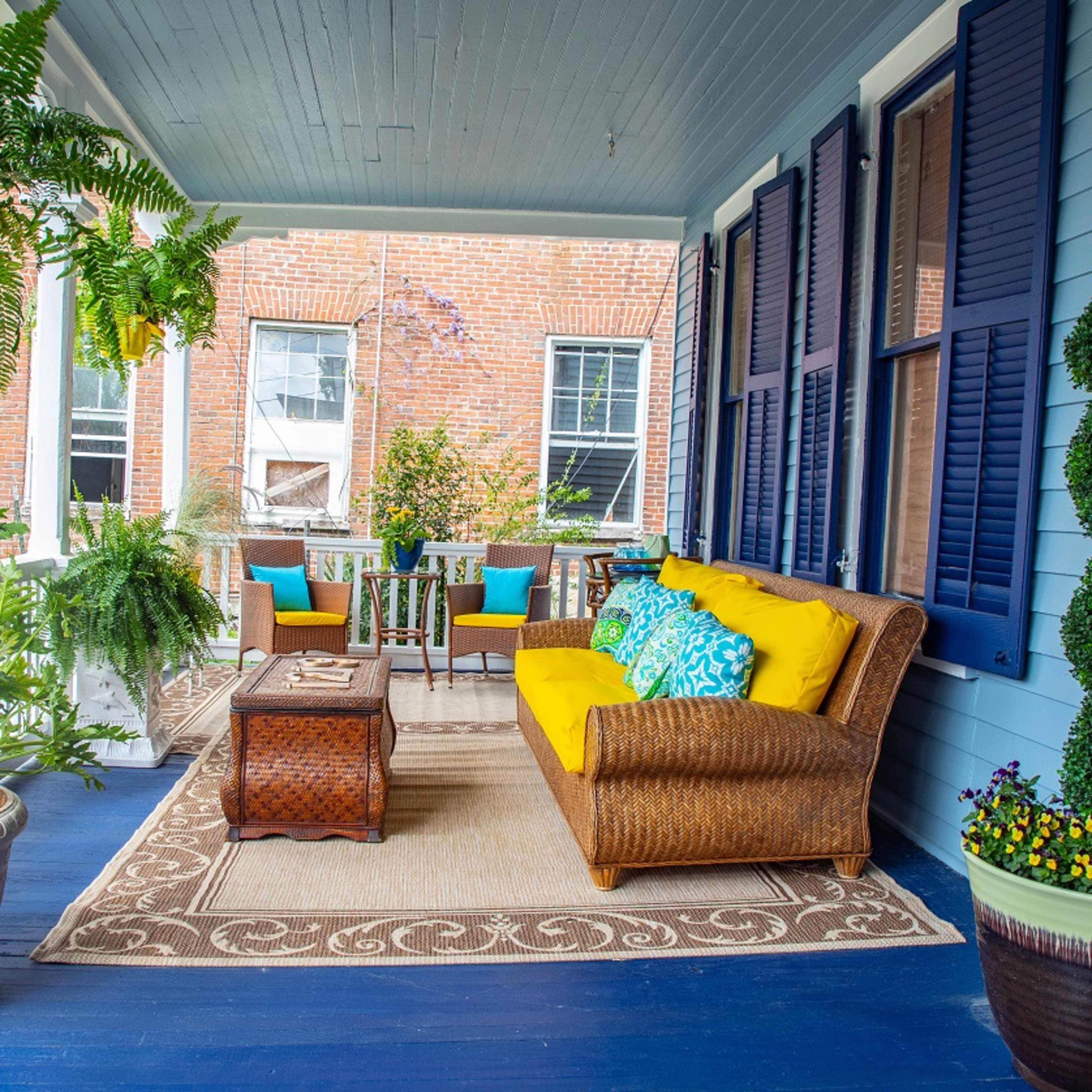 A covered porch features a wicker sofa with yellow cushions, a patterned area rug, potted plants, and a seating area with a wooden table surrounded by chairs.