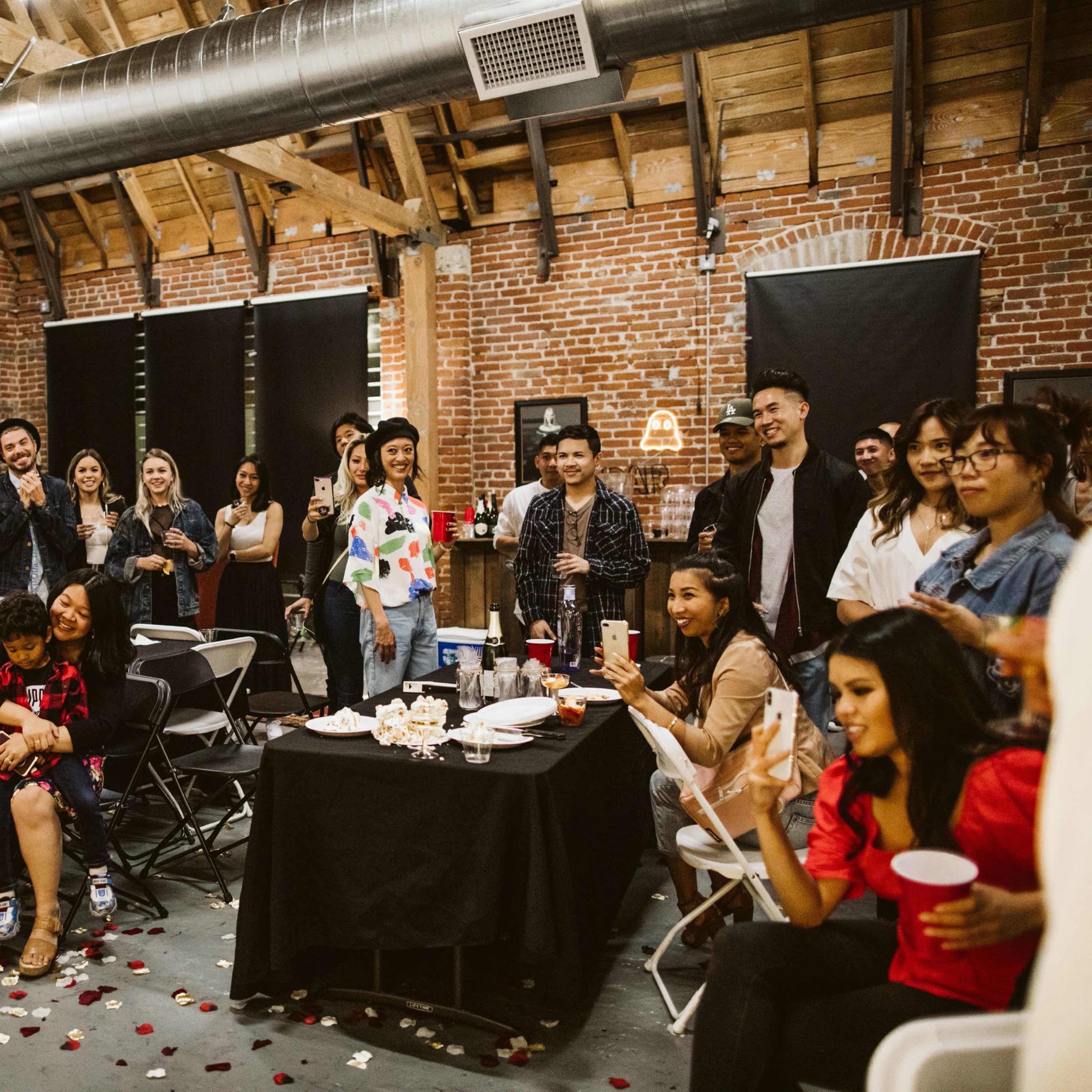 A group of people gathers in a brick-walled venue, some sitting at tables and others standing, while one person addresses the crowd.