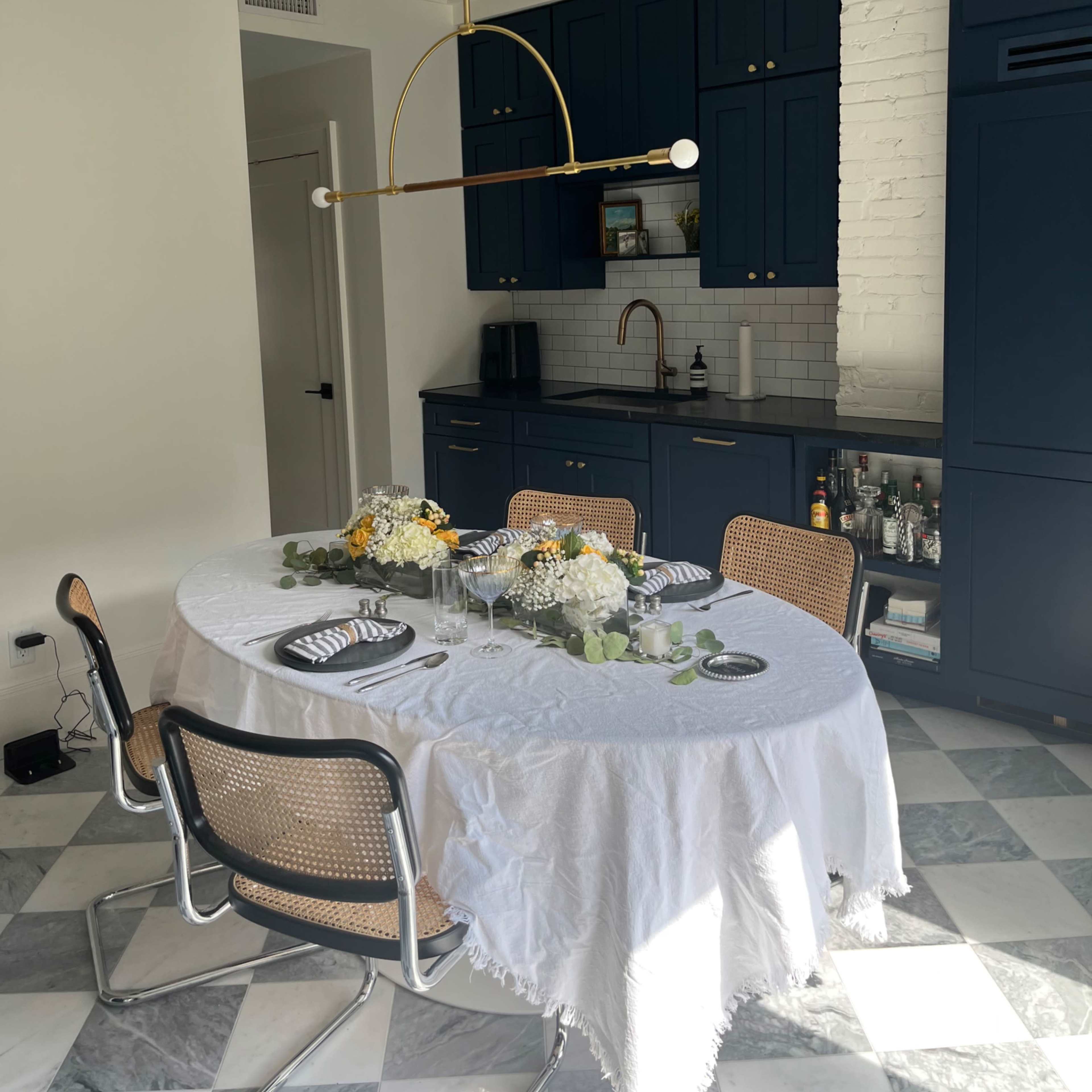 A modern kitchen with a round dining table set with plates and flowers, surrounded by chairs, and blue cabinetry visible in the background.