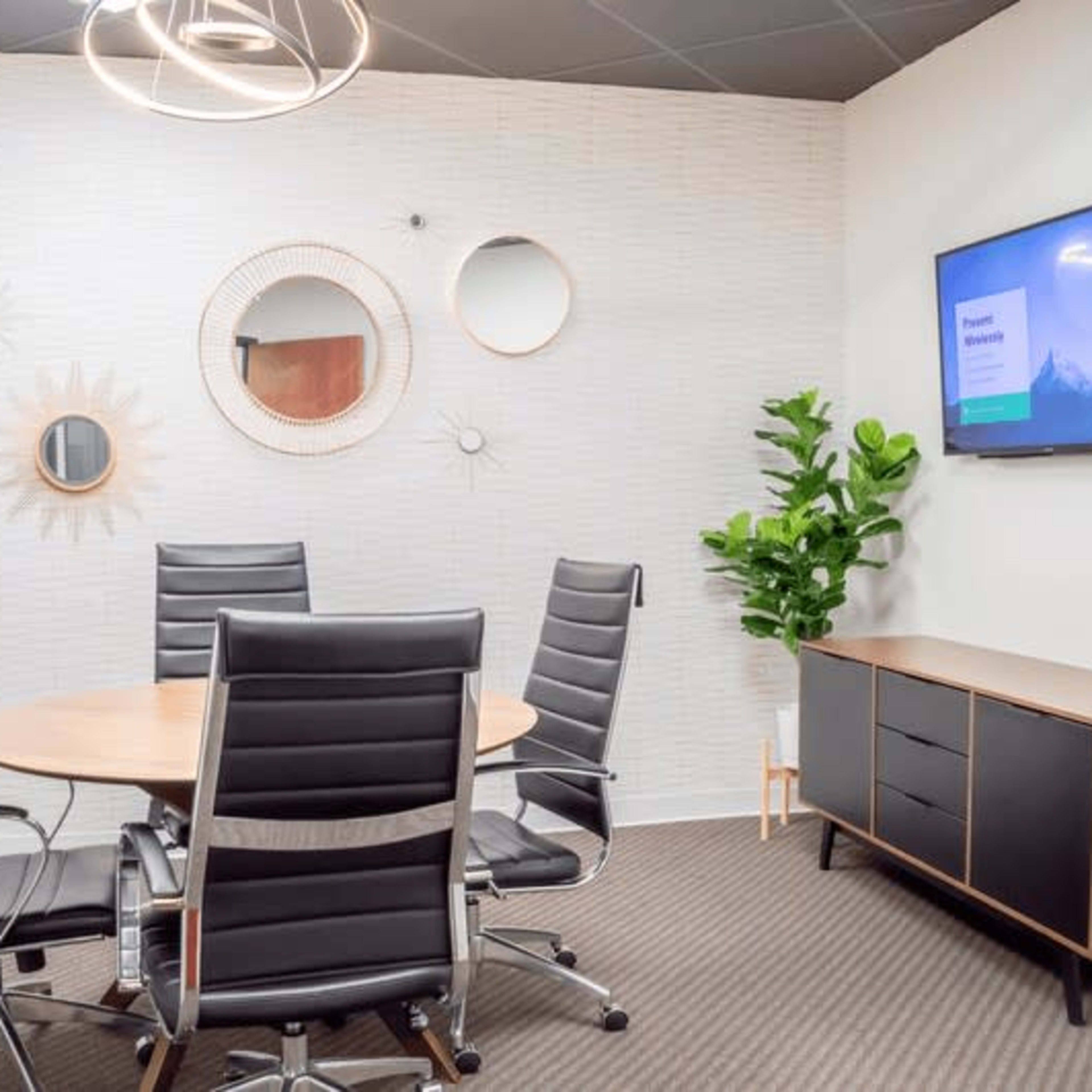 A small conference room with a round table surrounded by six black chairs, a plant, a wall-mounted TV, and decorative mirrors on the walls.