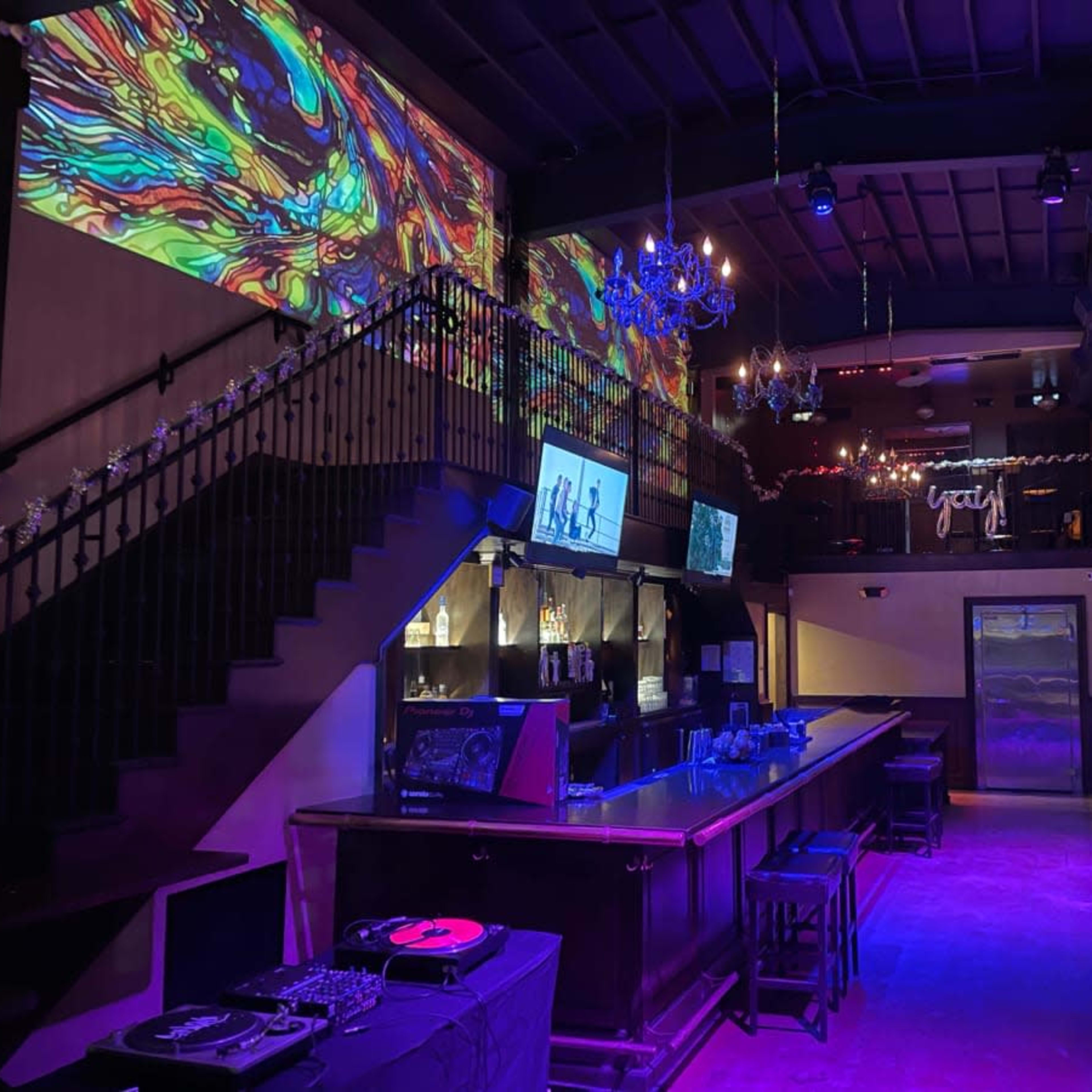 A dimly lit bar area with a staircase, colorful wall projections, and a DJ setup near the bar.