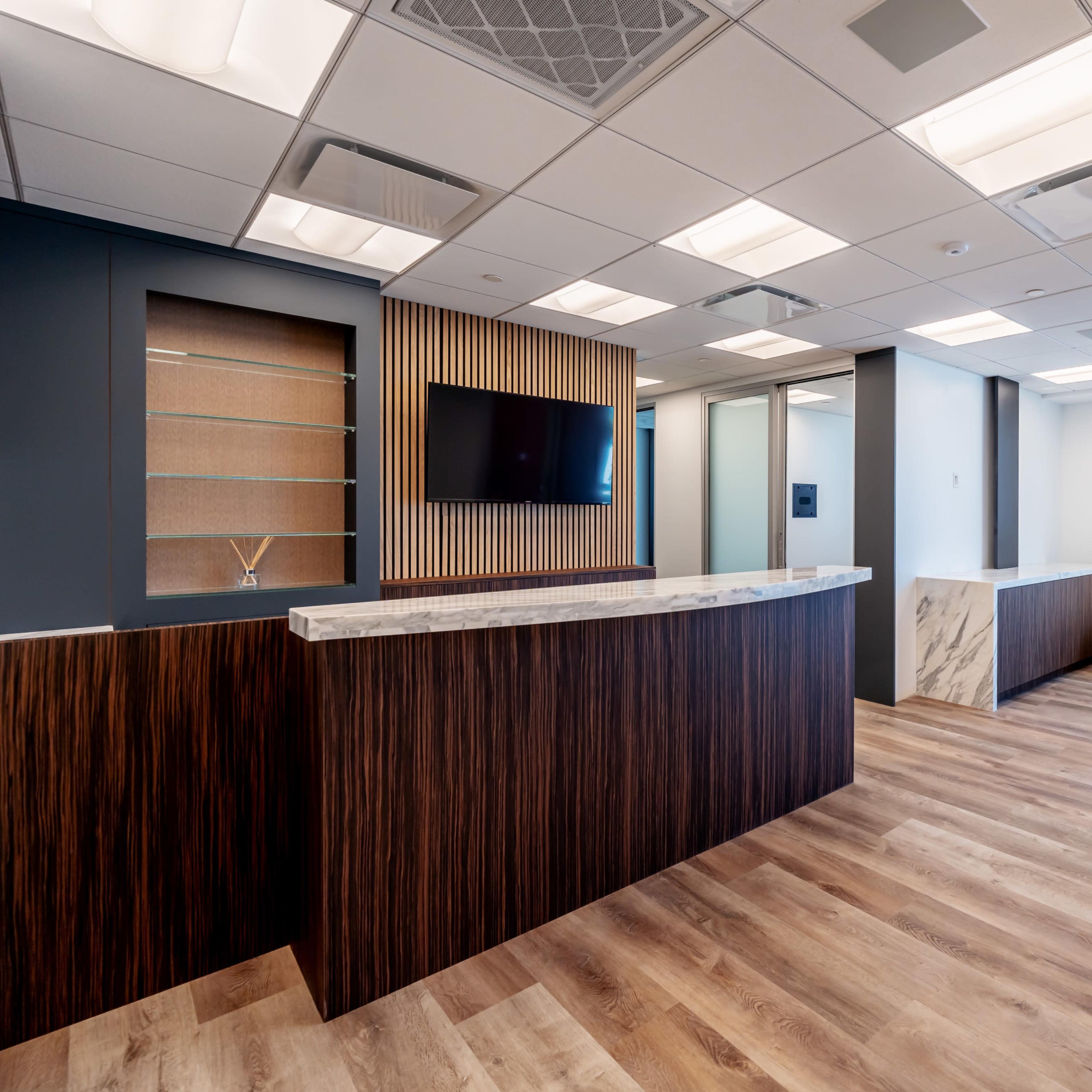 A modern office reception area with a dark wood reception desk, a wall-mounted TV, and a white sofa against a light-colored wall.