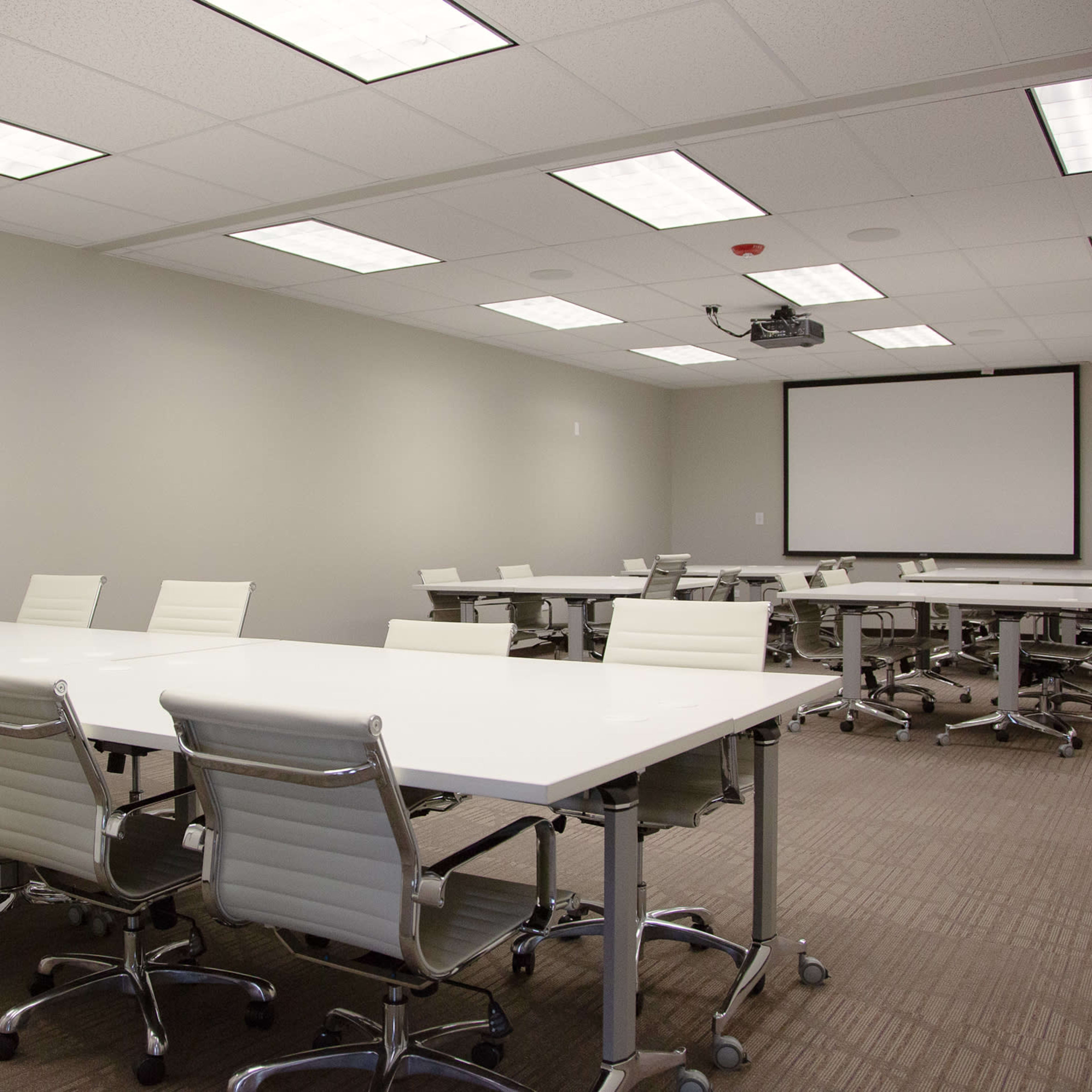 A modern meeting room with multiple white tables and chairs arranged in a symmetrical layout, equipped with a projector and two screens mounted on the walls.
