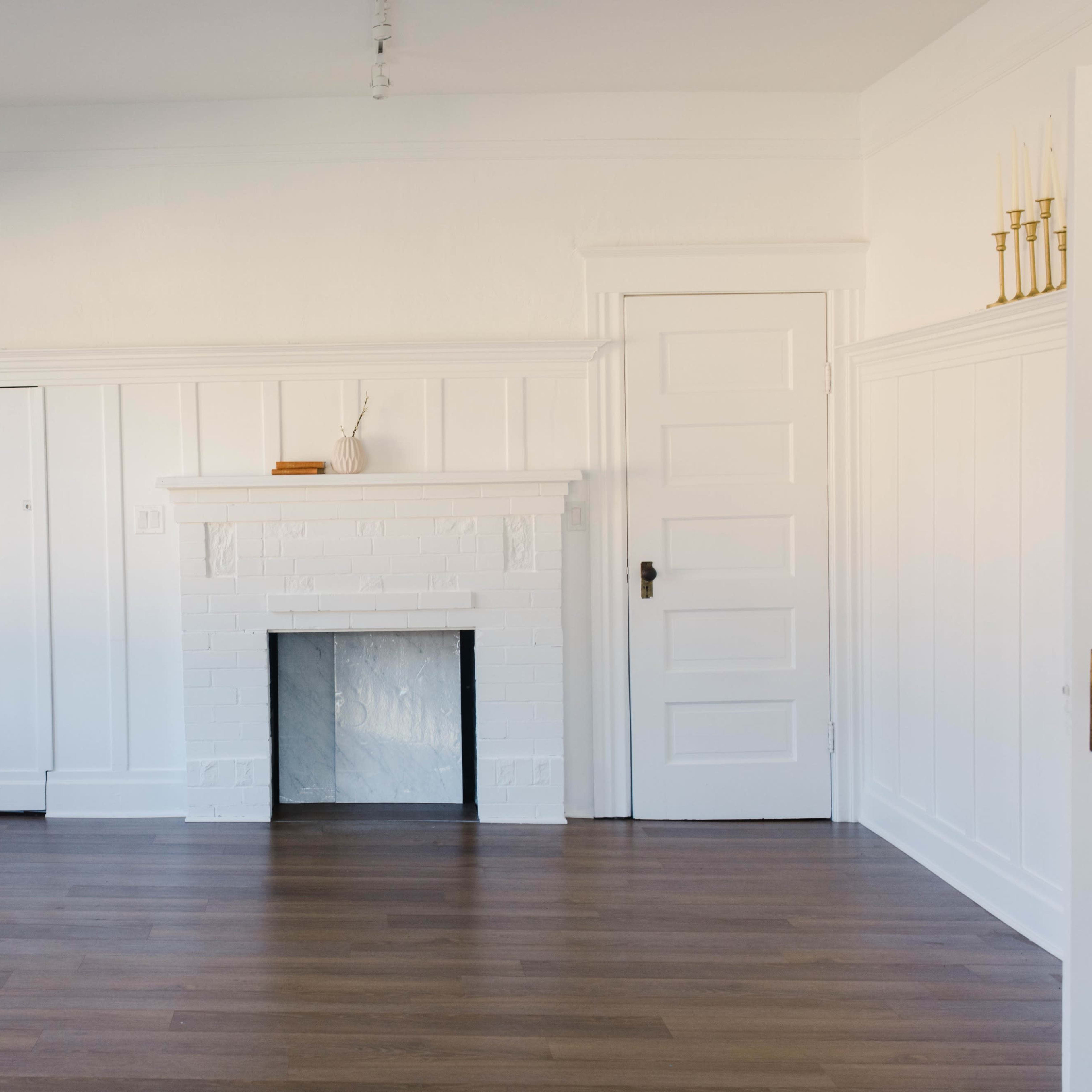 A bright, empty room with white walls, a brick fireplace, and wooden flooring.