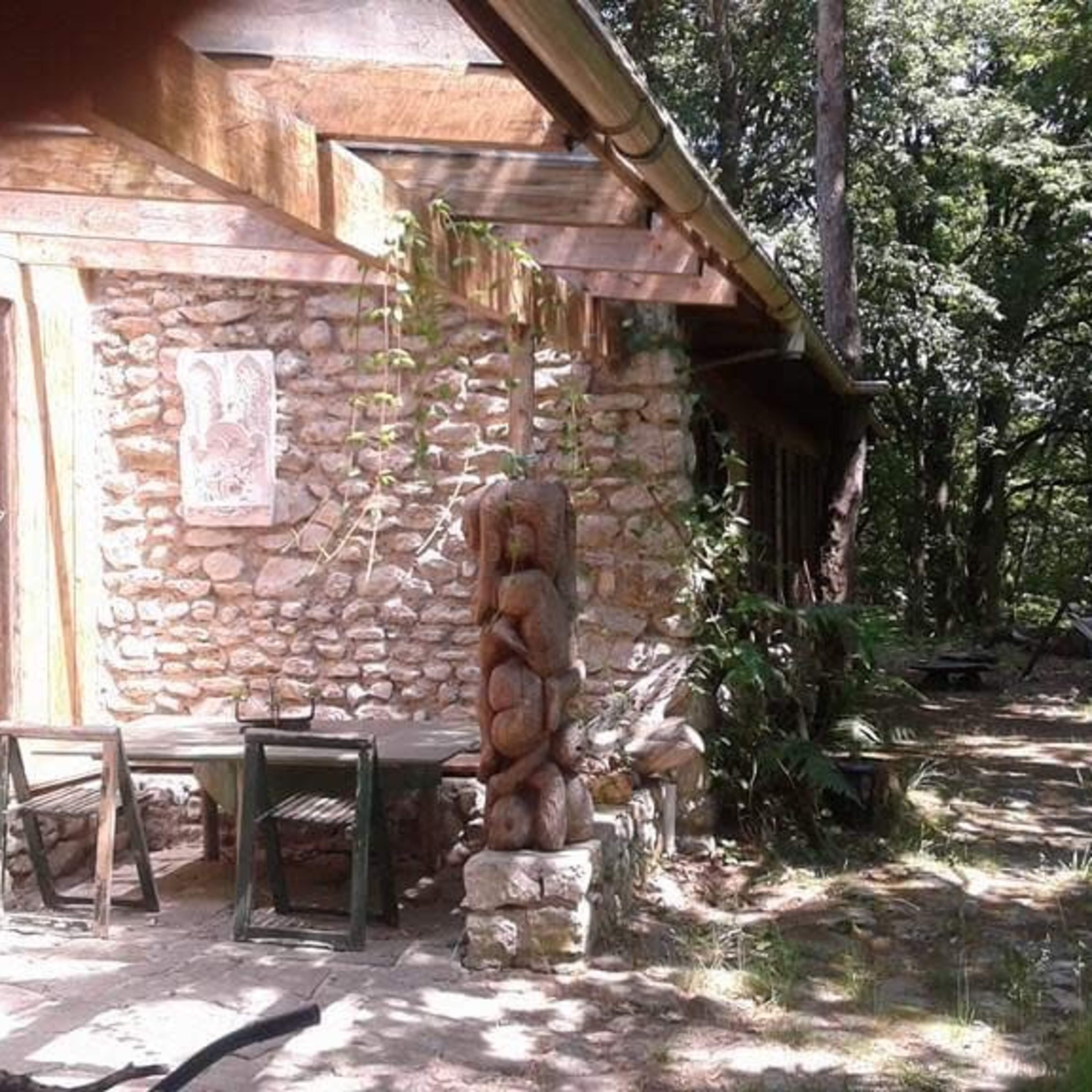 A stone and wood cabin is surrounded by trees, featuring a carved wooden sculpture and a table set on the patio.