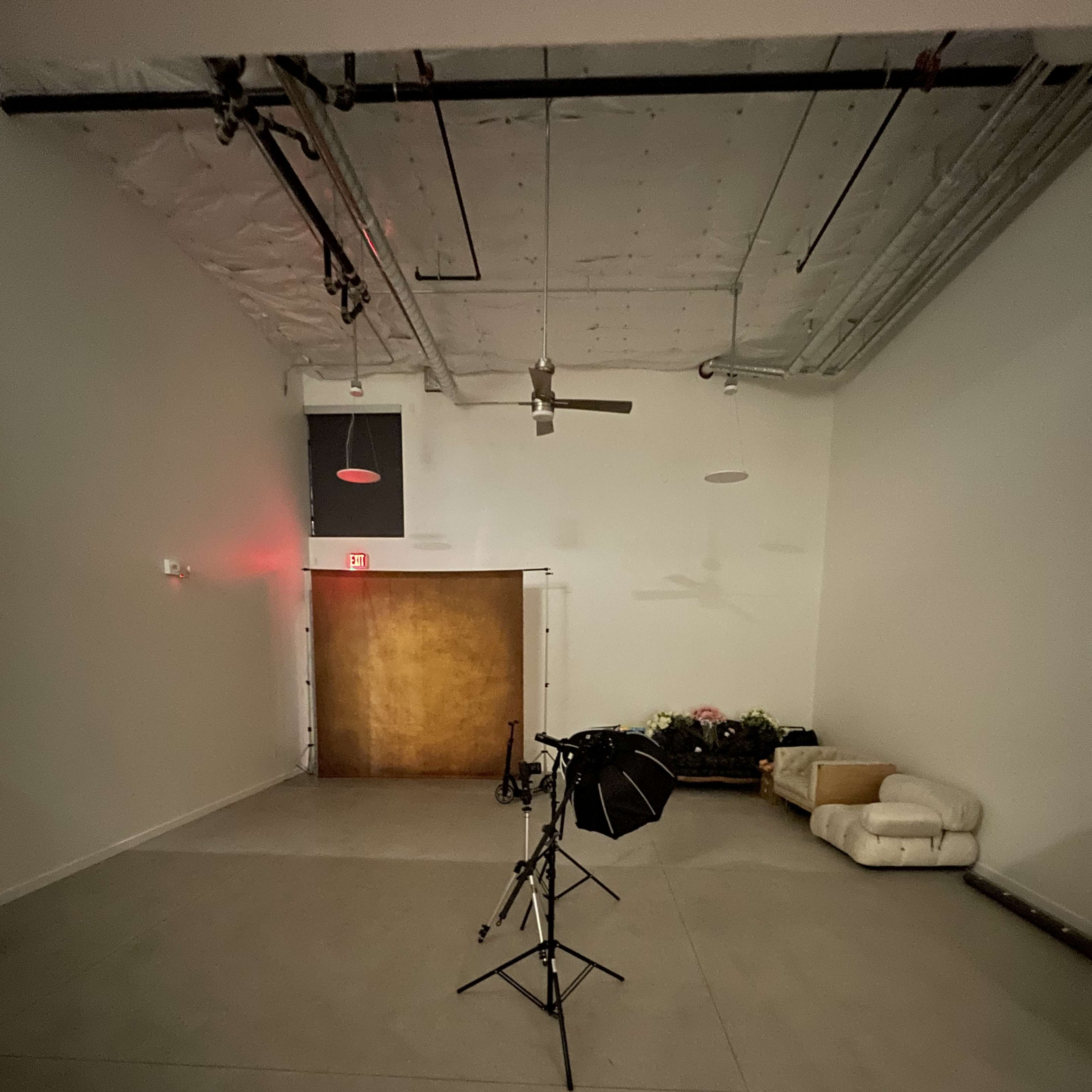 A spacious, empty room with a brown door at the far end, a soft white couch on the right, and a lighting stand positioned centrally in front.