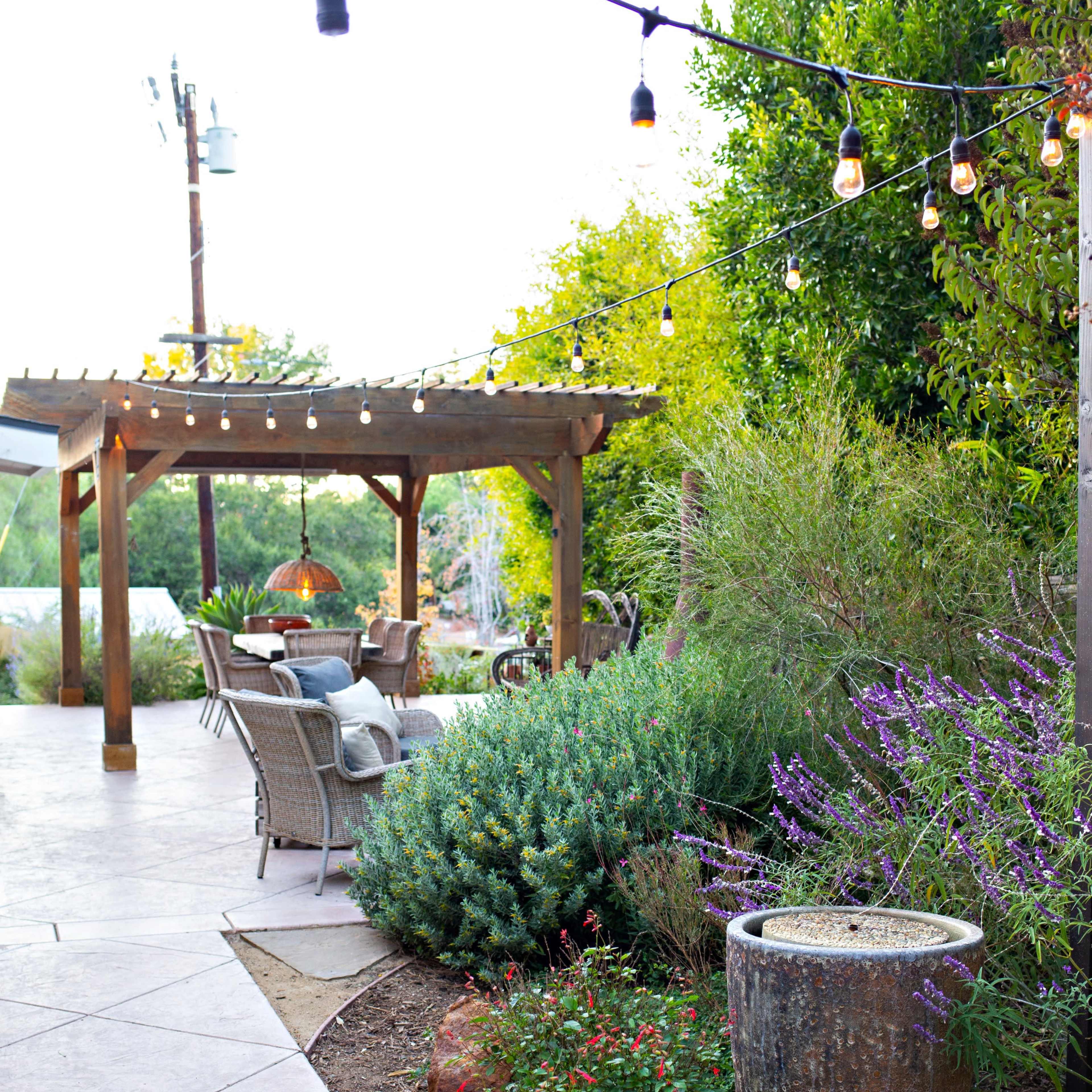 A landscaped outdoor area featuring a stone patio, a wooden pergola with string lights, and a variety of plants and seating.