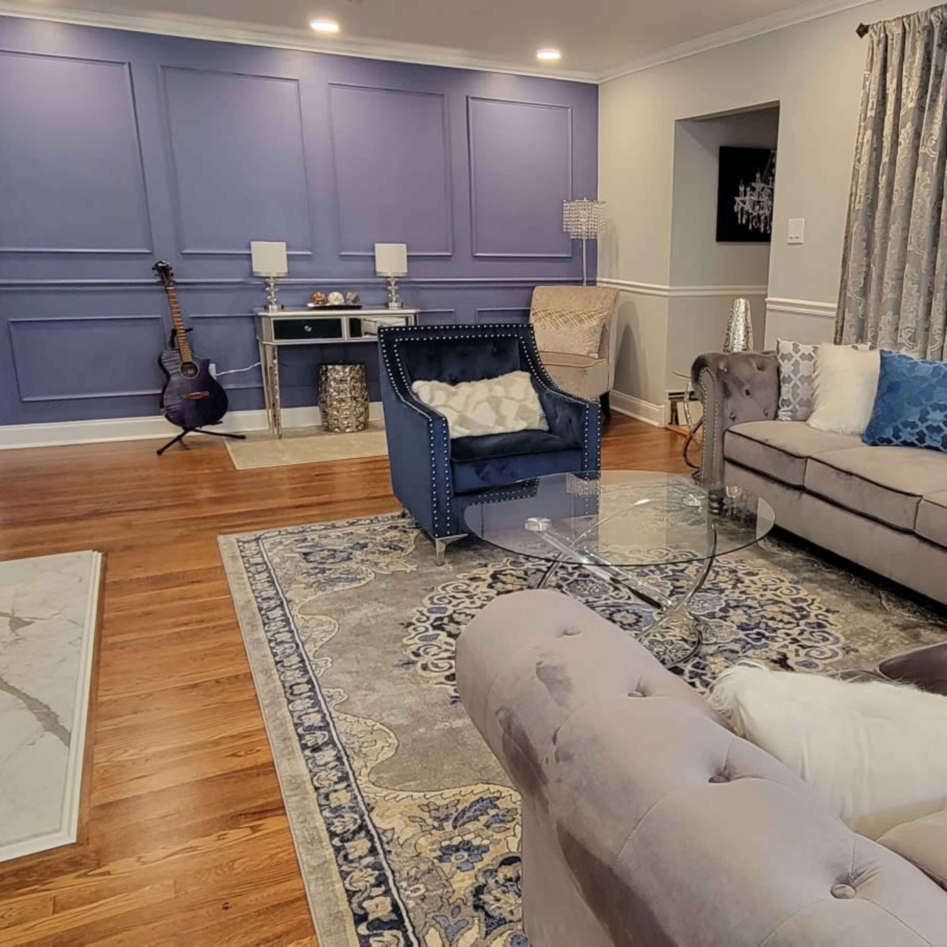 The living room features a light-colored sofa and armchairs arranged around a glass coffee table, with a rug on wooden floors and a blue accent wall.