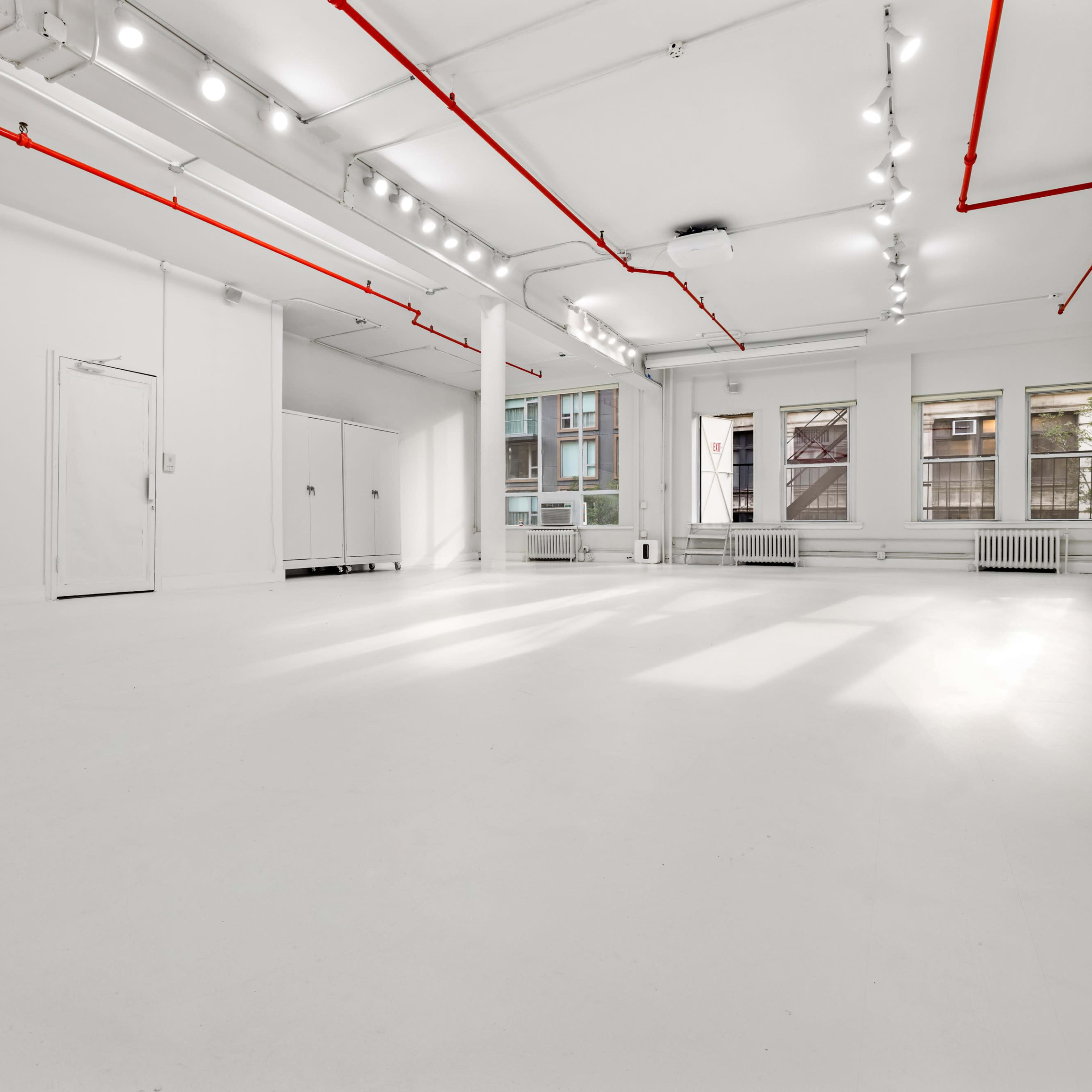 A spacious, empty room with white walls, a white floor, and red pipes along the ceiling.