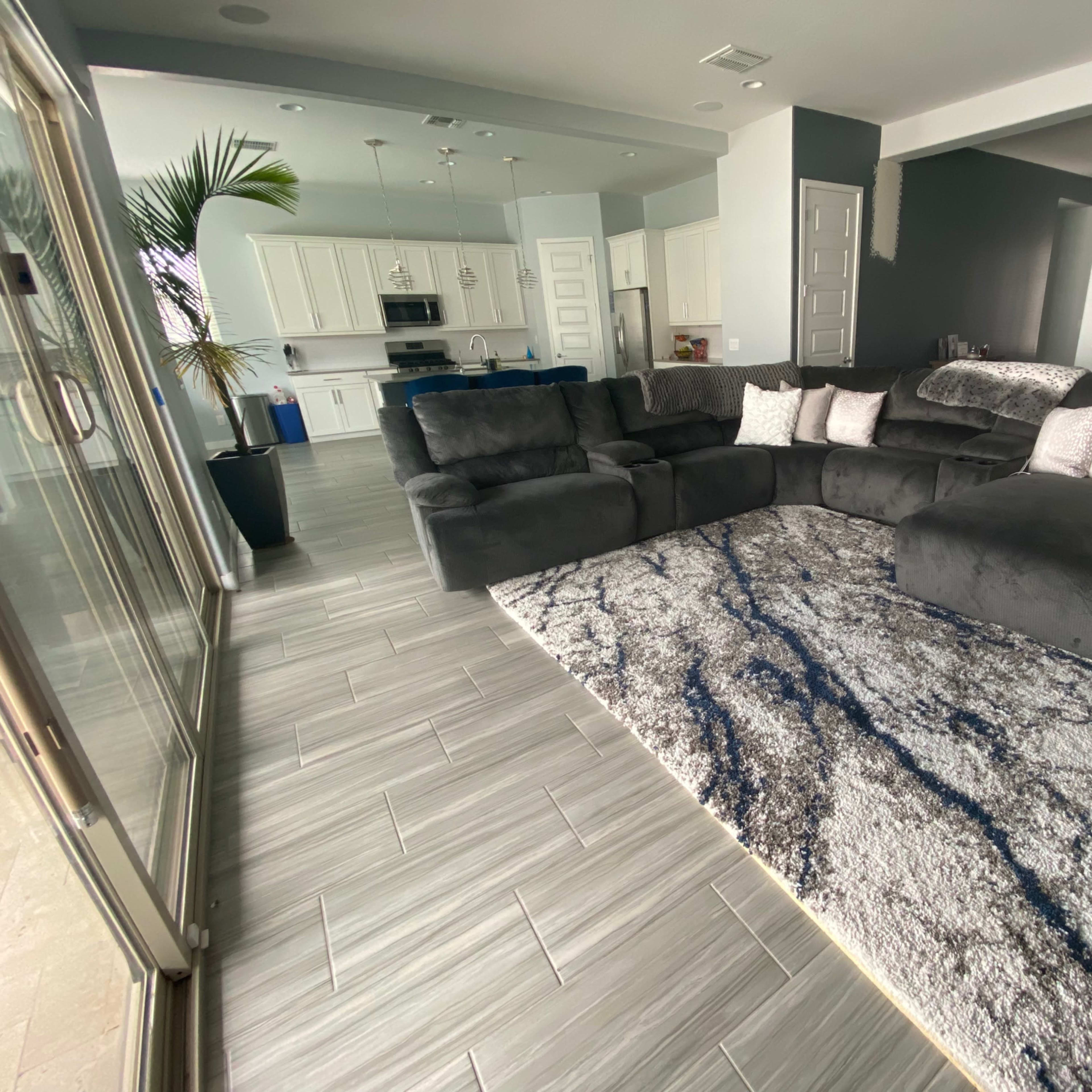 A modern living room features a gray sectional sofa, a patterned area rug, and framed photographs on the wall, with an open kitchen visible in the background.