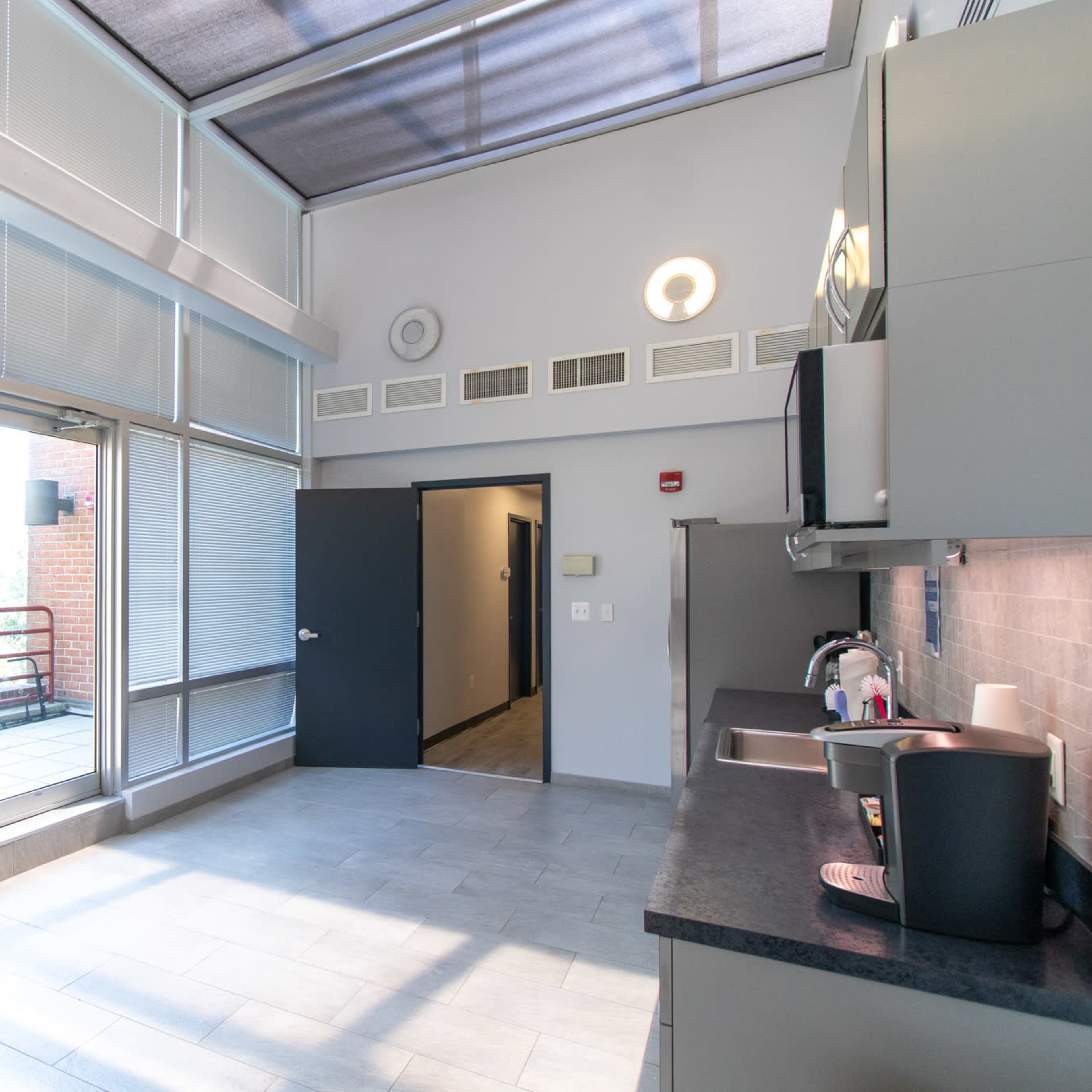 A modern kitchen space with large windows, a door leading to a balcony, and minimalist cabinetry and appliances.
