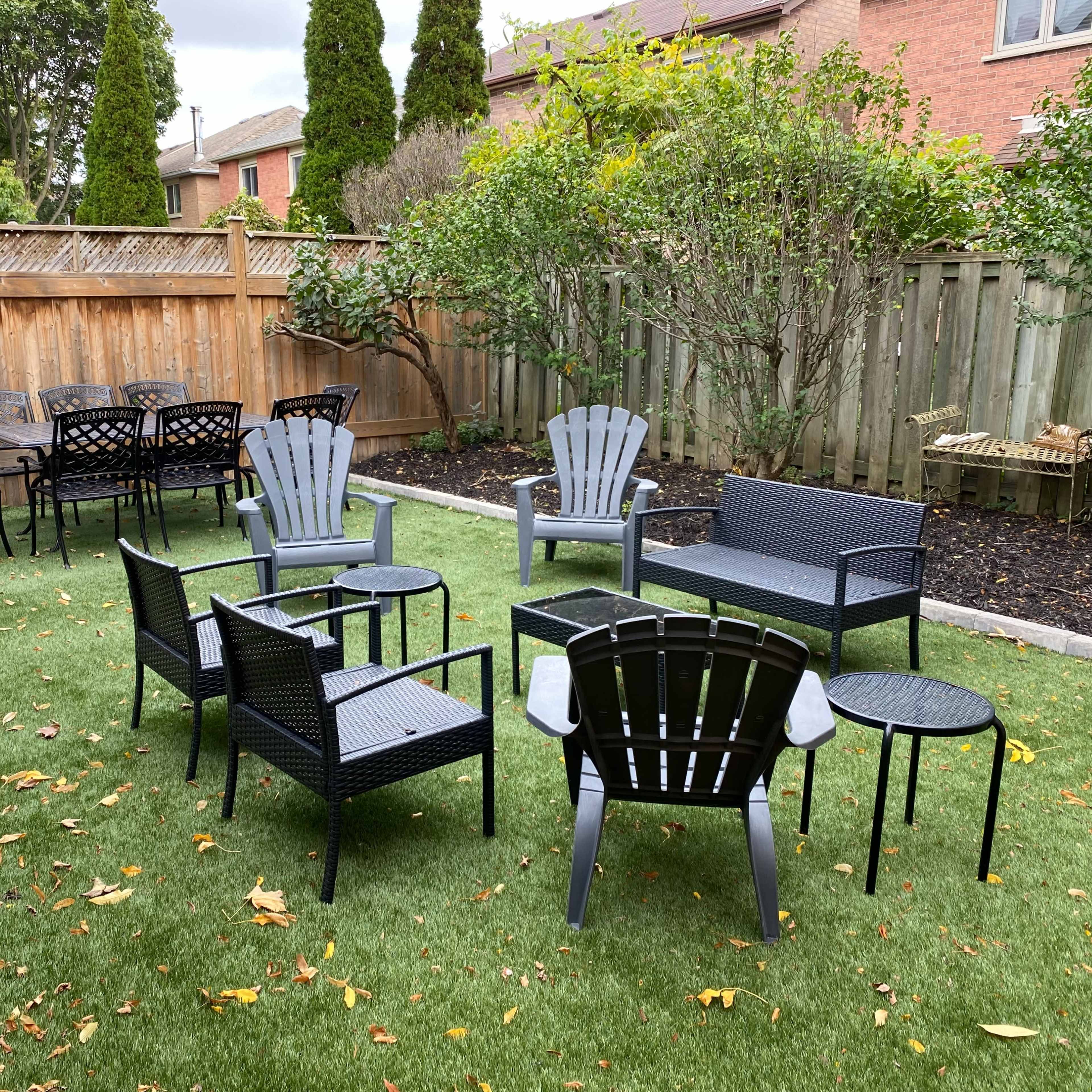 A landscaped backyard with several outdoor seating sets arranged on a patch of artificial grass, surrounded by wooden fencing and small trees.