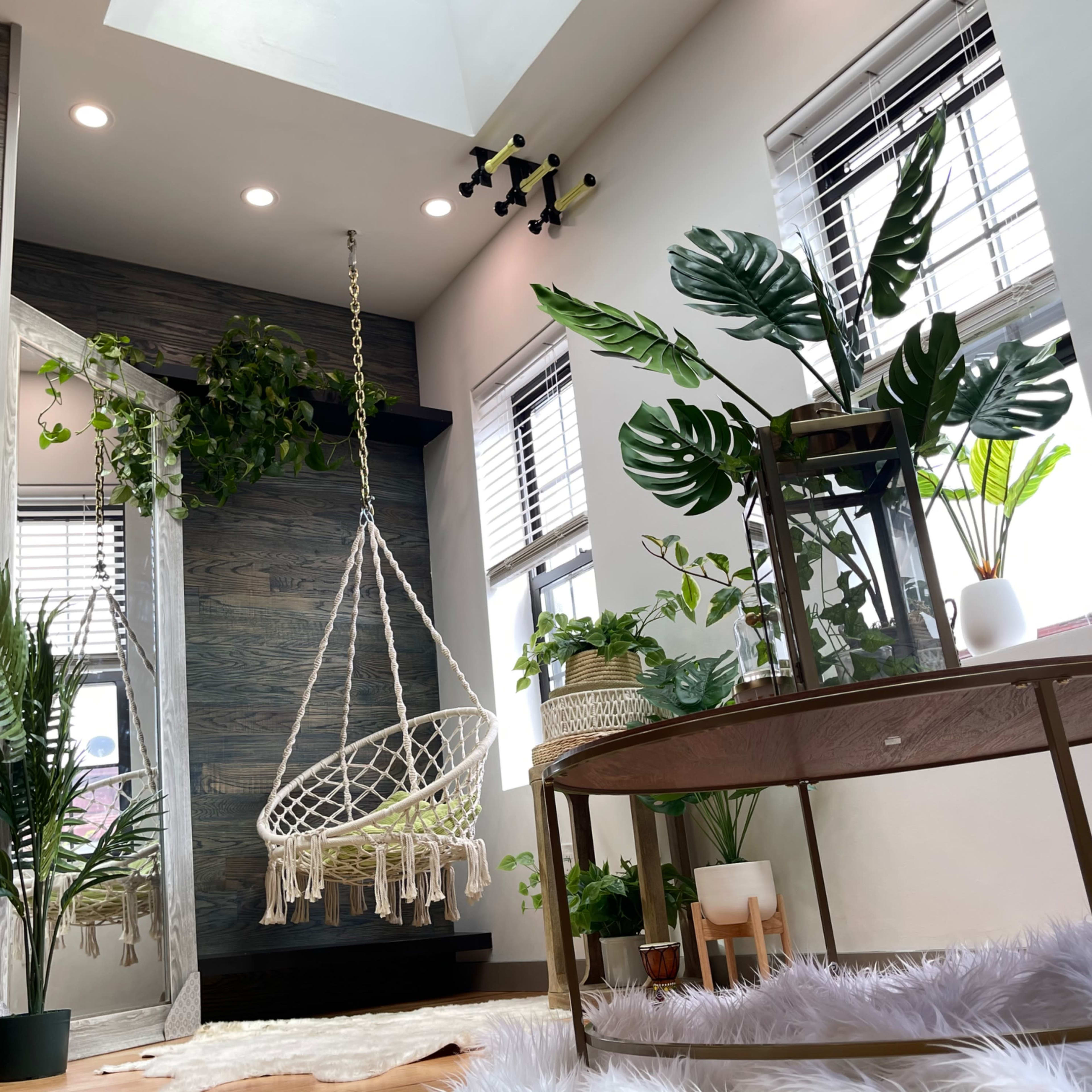 A cozy indoor space featuring a hanging macramé chair, various potted plants, and a round coffee table with a decorative item on it.