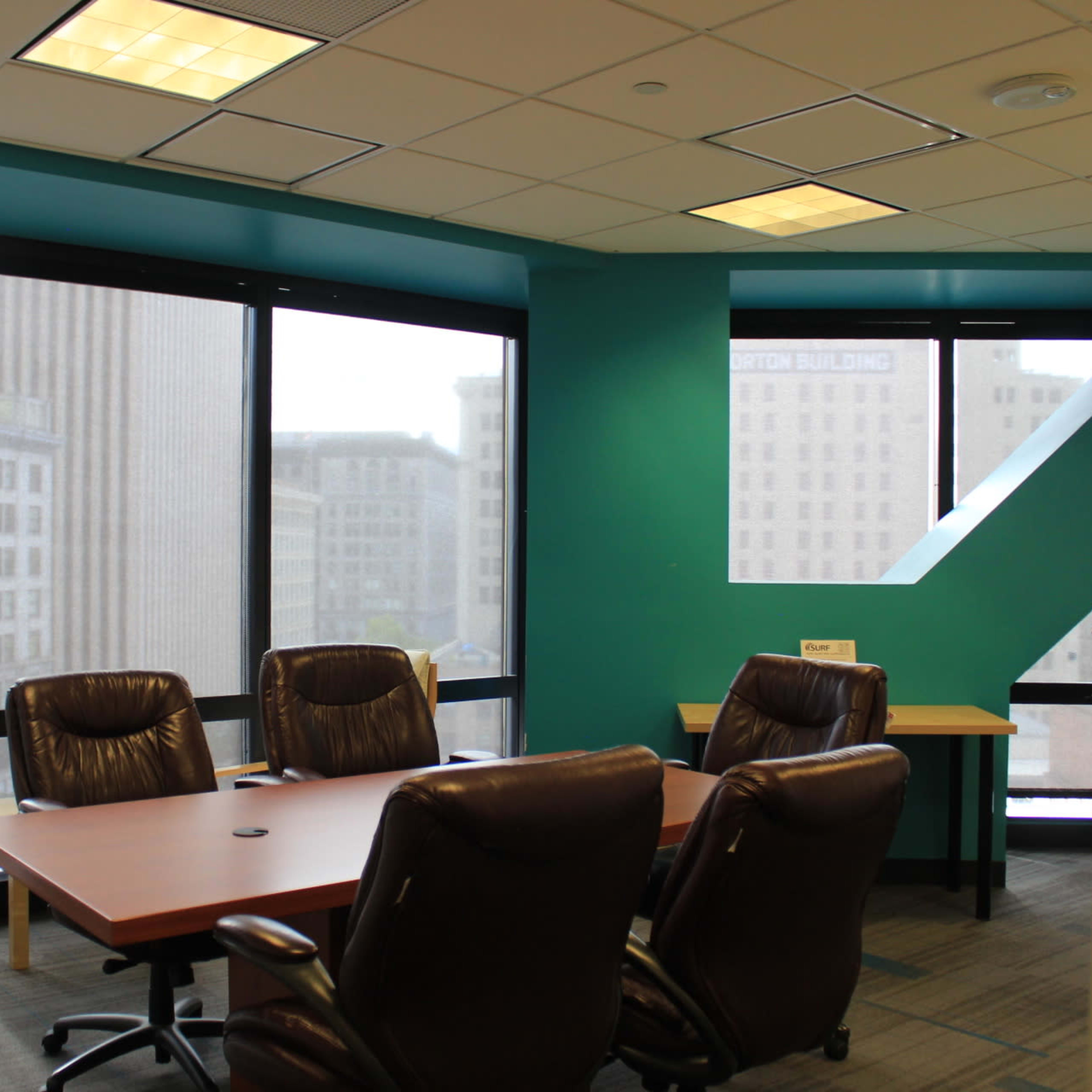 A conference room with a large table and several ergonomic chairs is illuminated by natural light from large windows overlooking a cityscape.