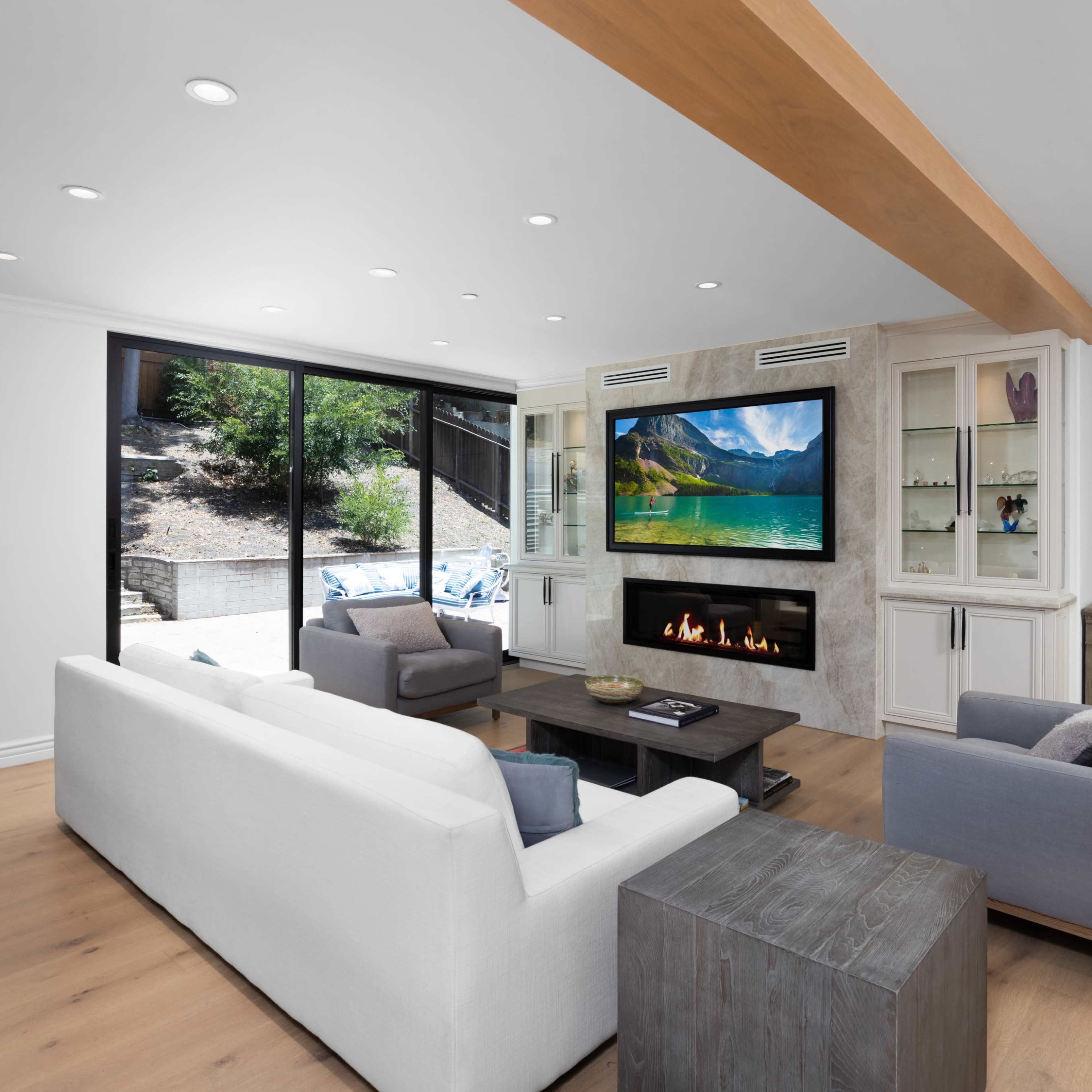 The living room features white and gray sofas arranged around a coffee table, with a modern fireplace and a large television set into the wall, and large windows providing a view of the outdoors.