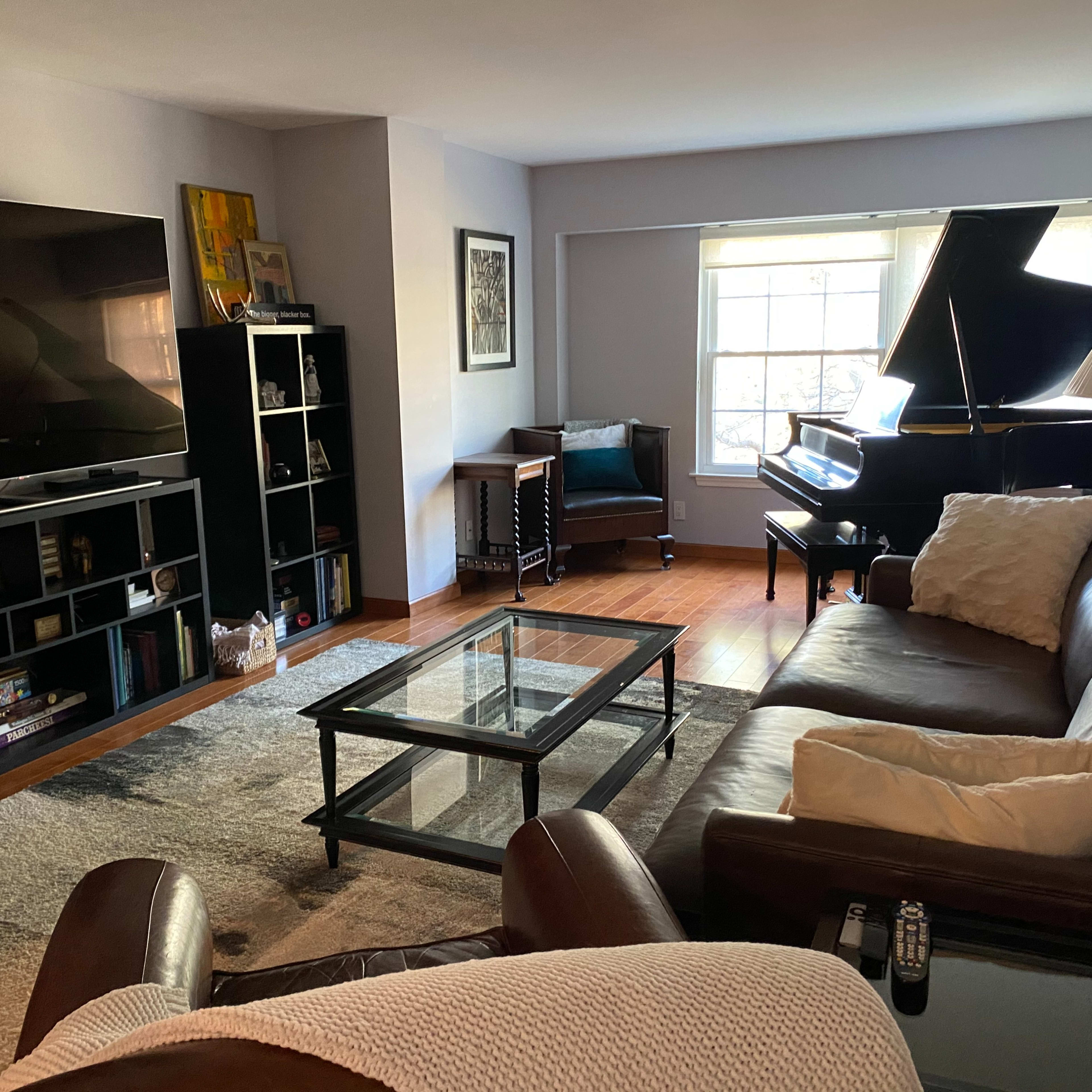 A living room features a large television, a glass coffee table, a leather sofa, a grand piano, and a cozy armchair by a window.