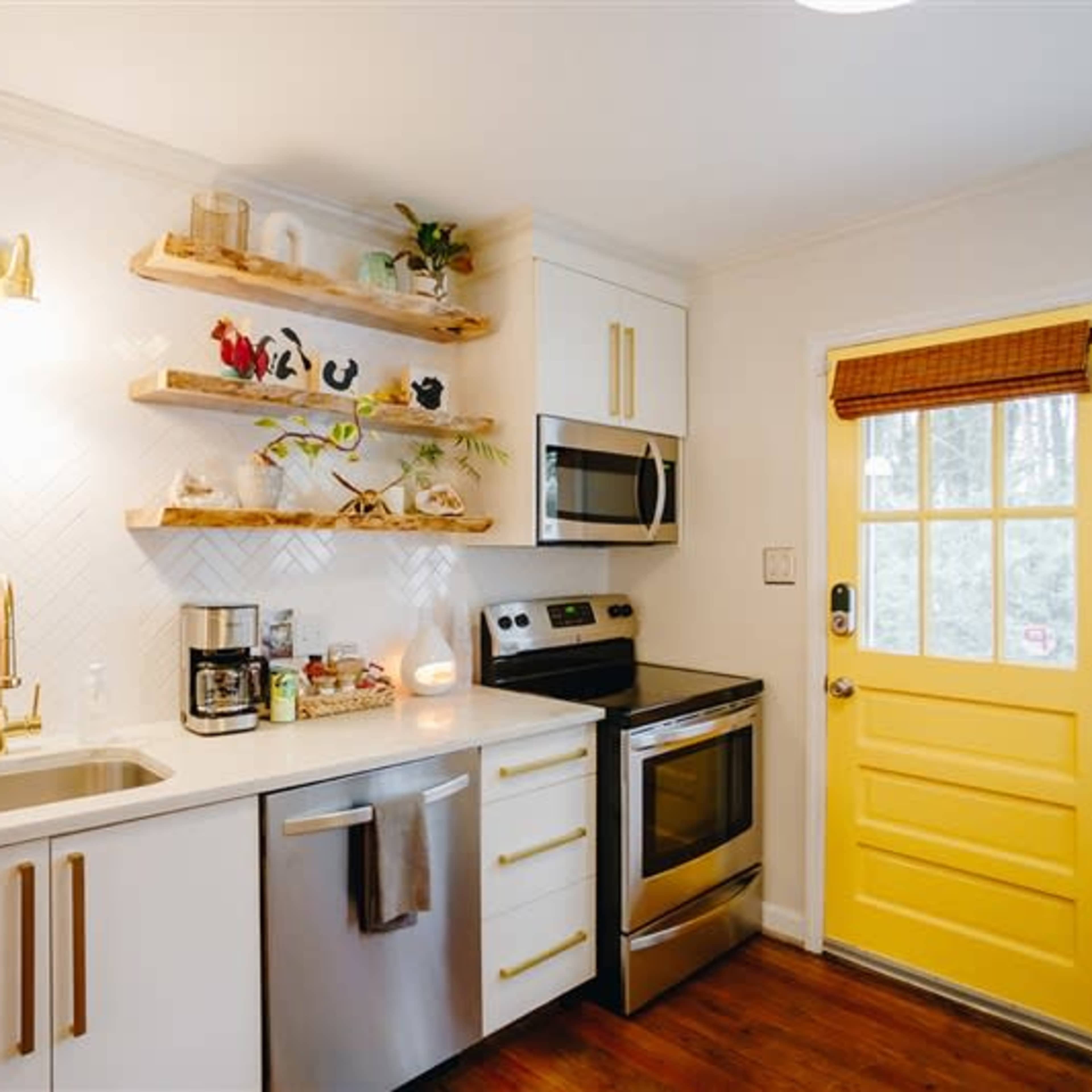 A small kitchen features open wooden shelves, stainless steel appliances, and a bright yellow door.