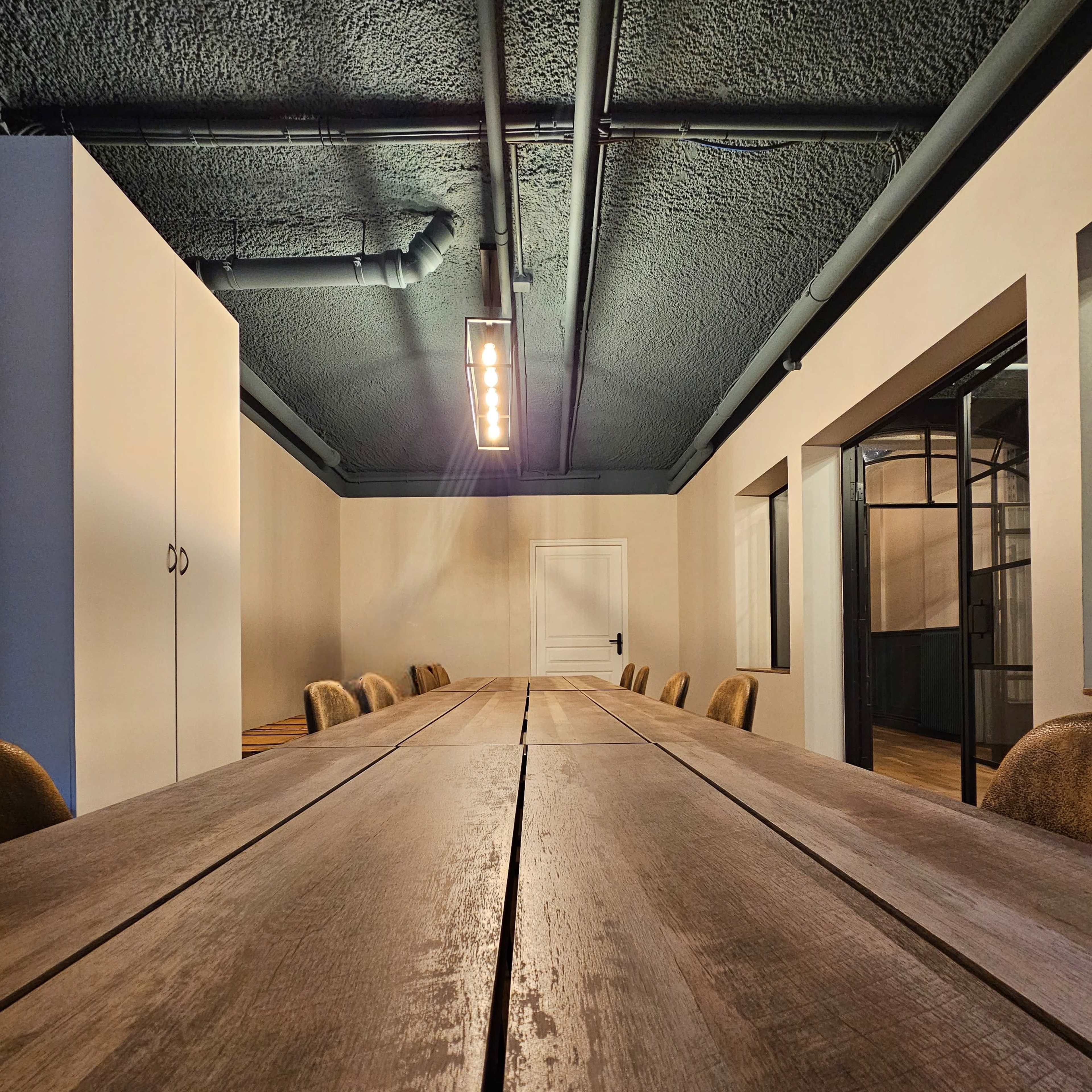 A long wooden table is centered in a spacious conference room with minimalist decor and industrial features.