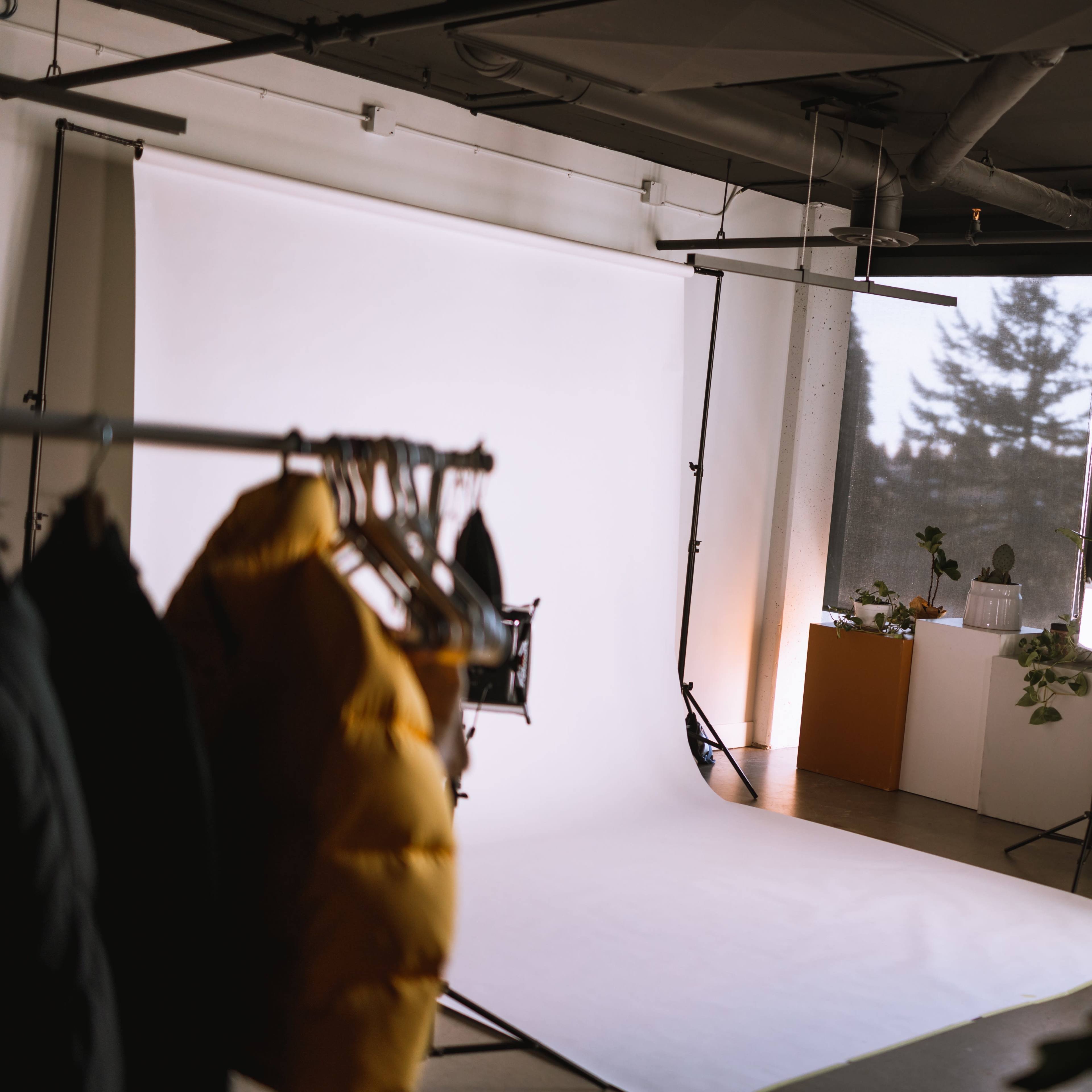 A well-lit photography studio features a white backdrop, clothing on a rack, and potted plants on display by a large window.