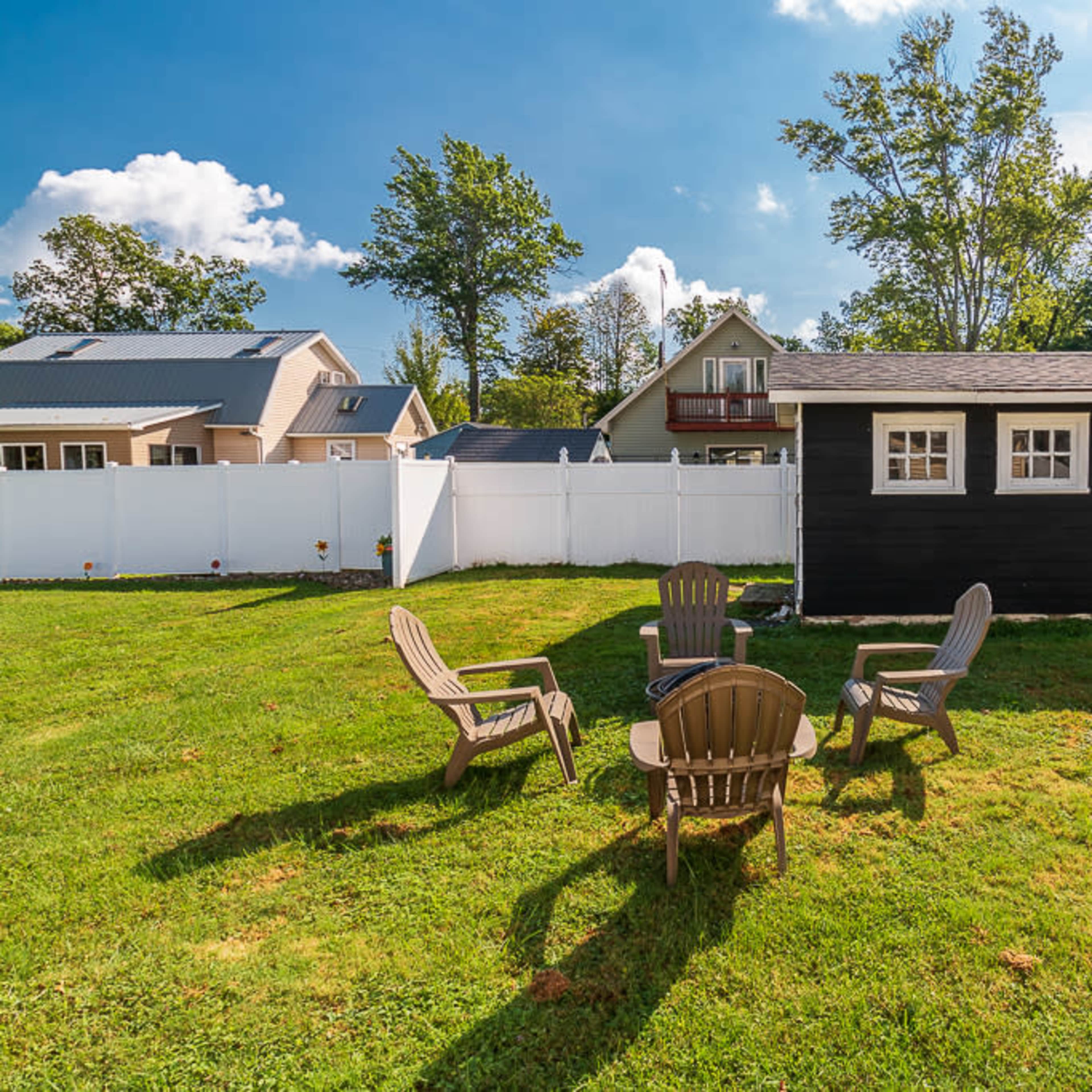 A small black shed in a grassy yard surrounded by a white fence, with four wooden patio chairs arranged in a circle nearby.