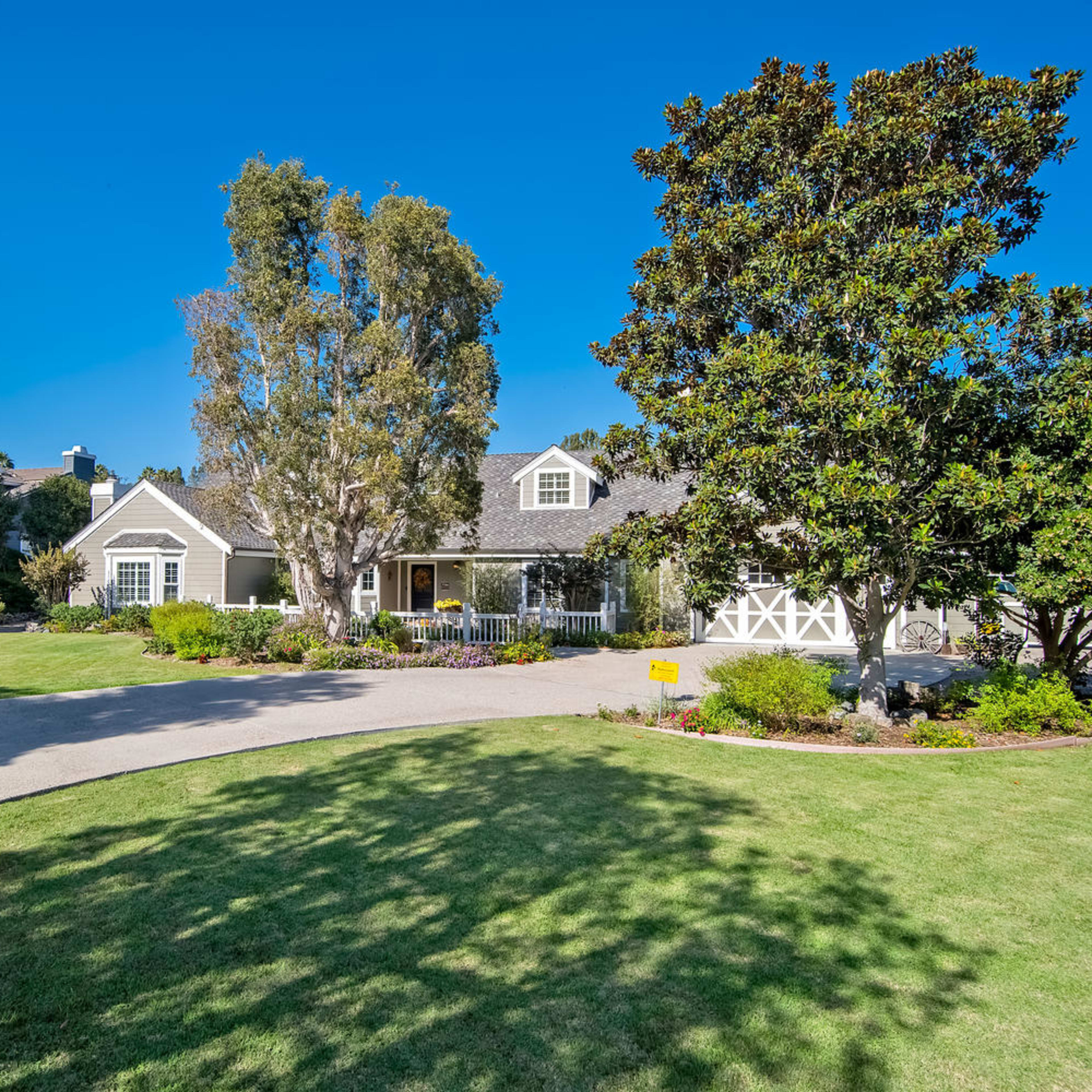A single-story house with a large front yard and driveway, framed by several trees and shrubs under a clear blue sky.