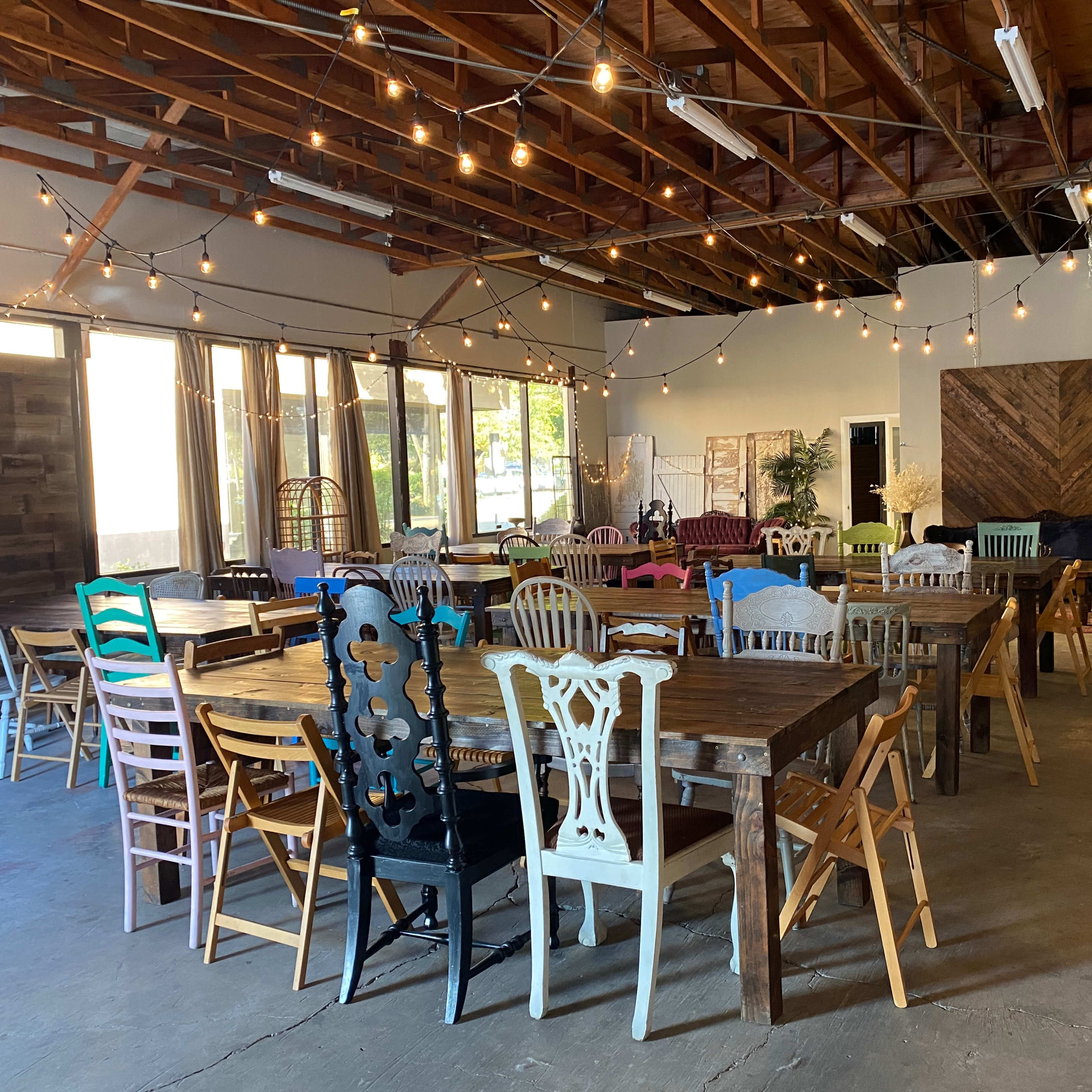 A spacious indoor gathering area filled with various wooden tables and colorful chairs, featuring string lights overhead and large windows letting in natural light.