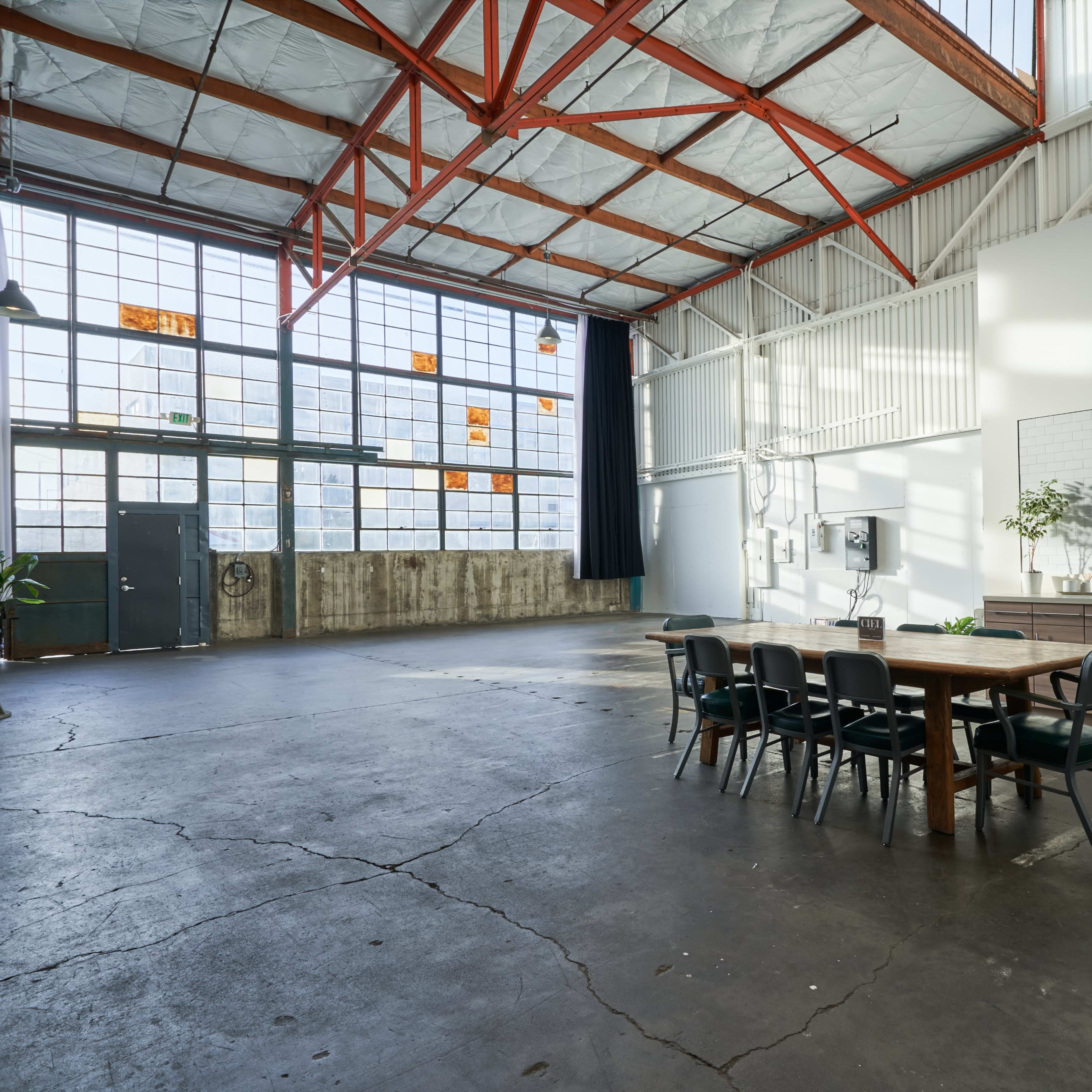 A spacious industrial-style interior with high ceilings, large windows, and a wooden dining table surrounded by black chairs, adjacent to a kitchen area.