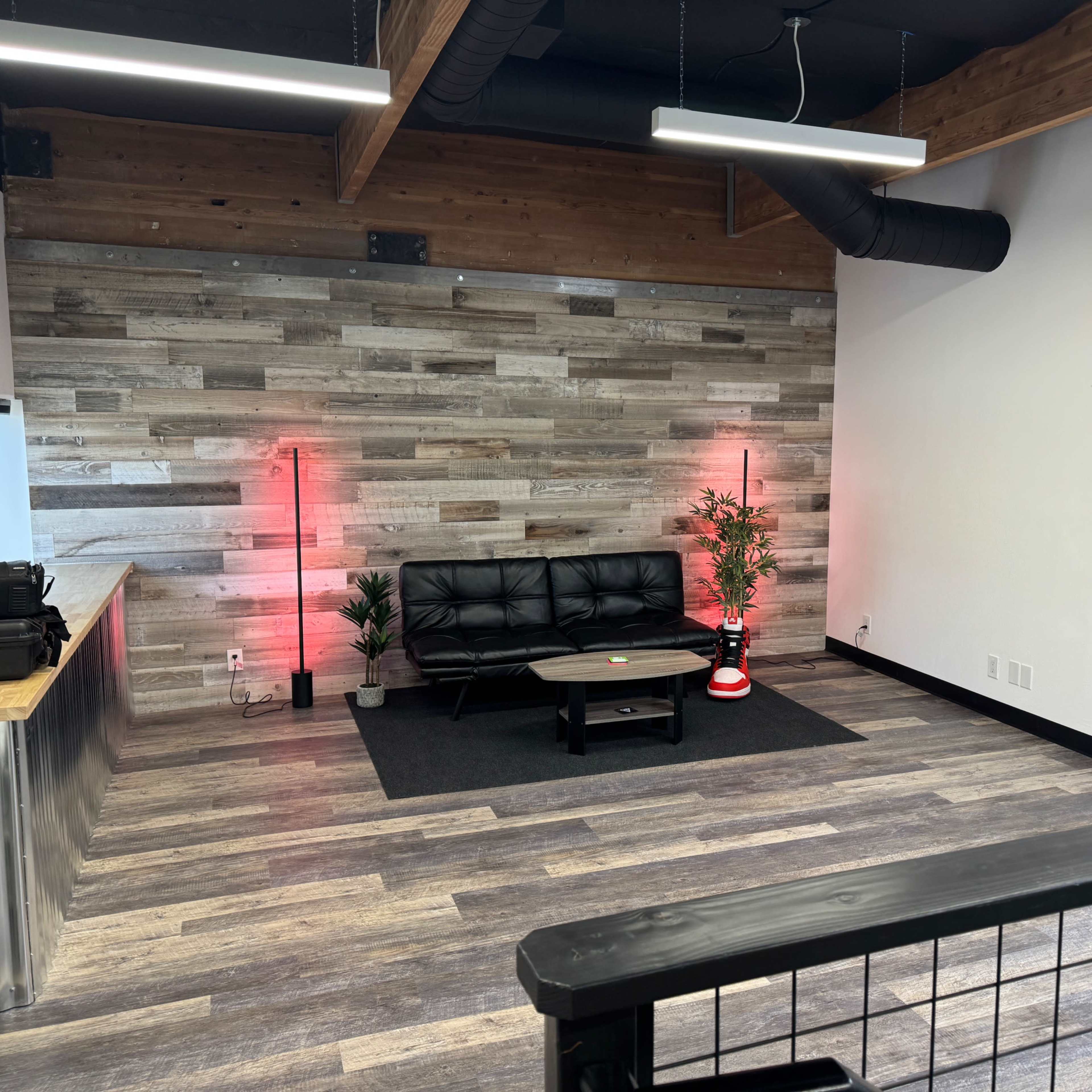 A modern office space with a black couch, a wooden accent wall, and plants, illuminated by red light lamps.
