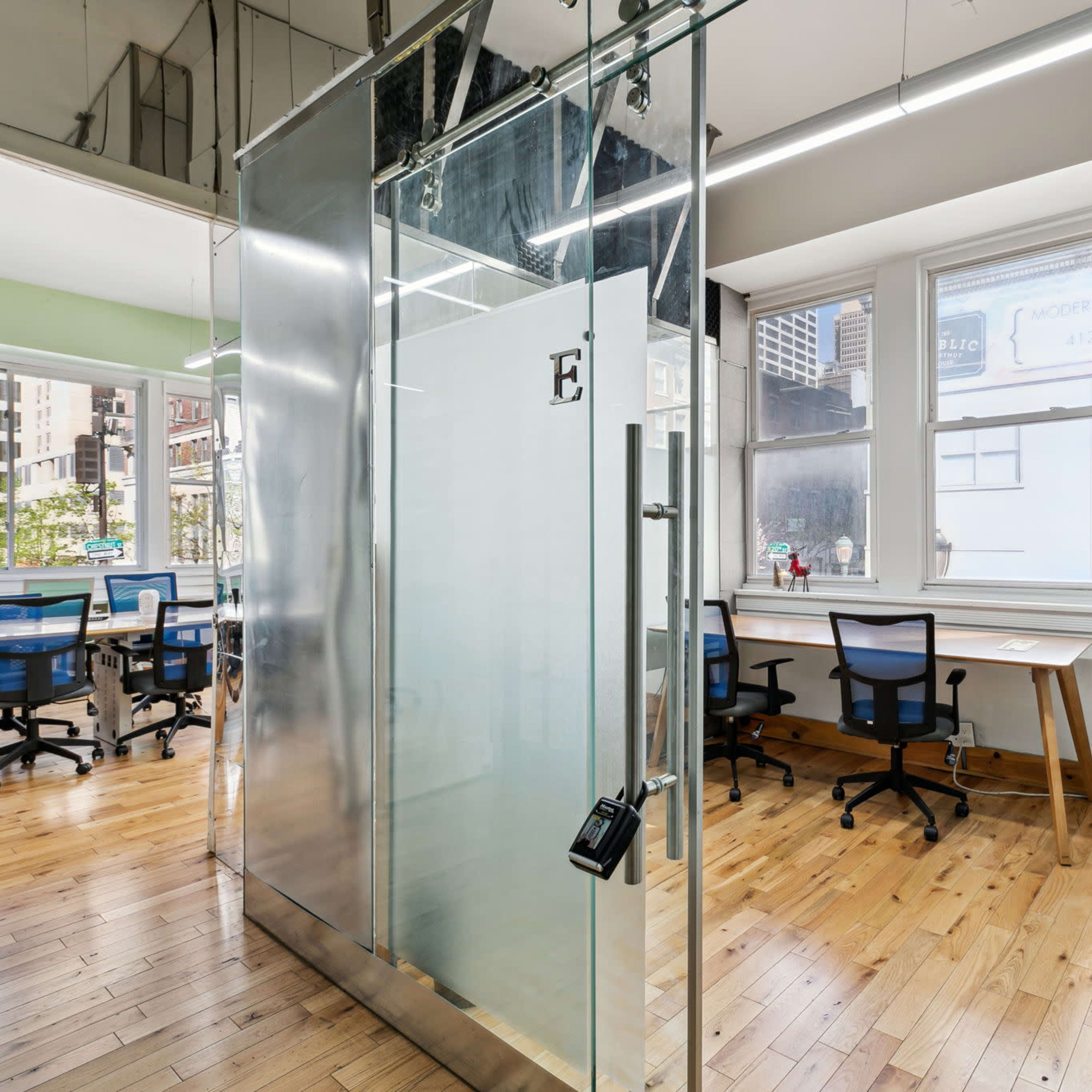 A modern office space featuring glass meeting rooms, wooden flooring, and several workstations with chairs.