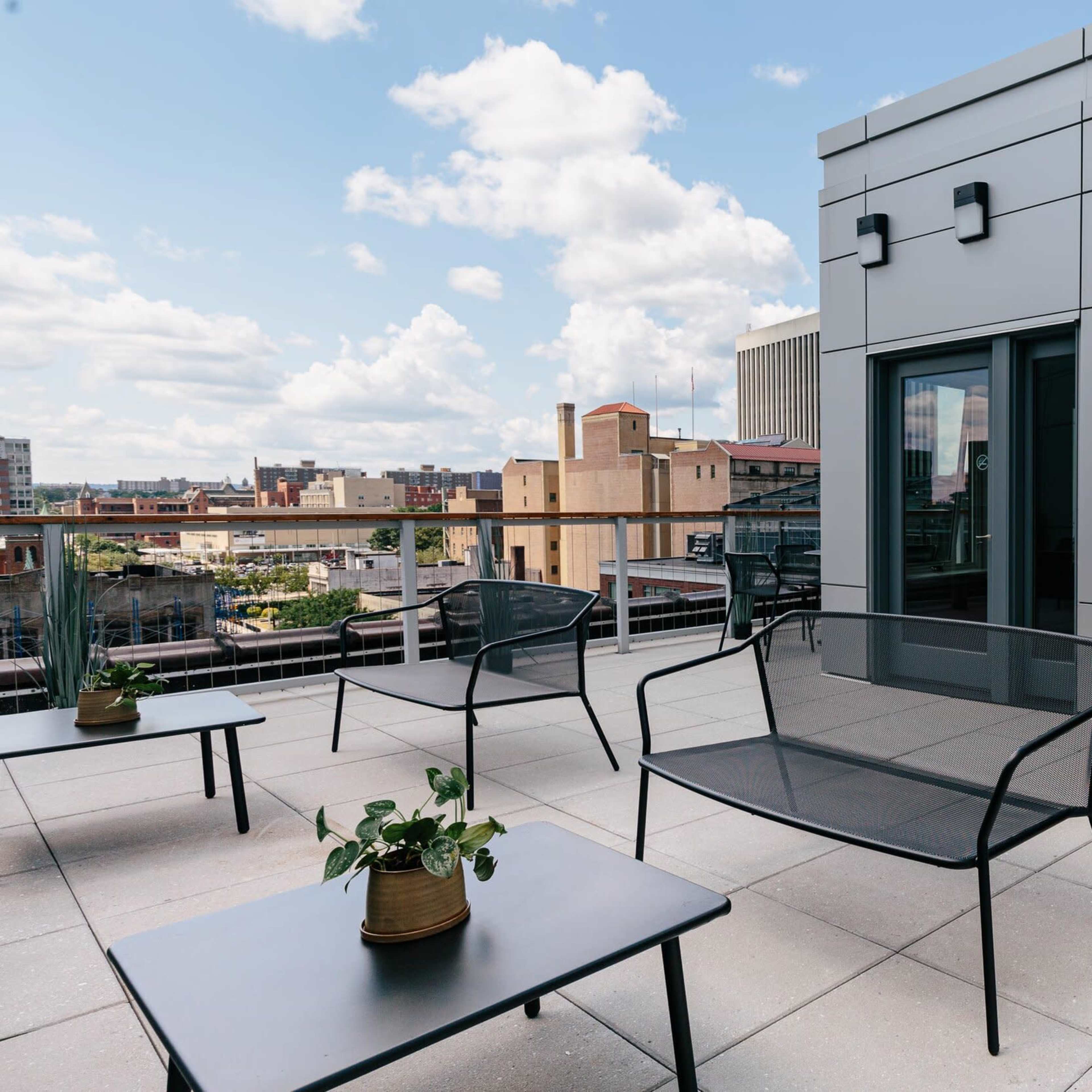 A rooftop terrace featuring a grill, black metal seating, and a view of the city skyline under a partly cloudy sky.