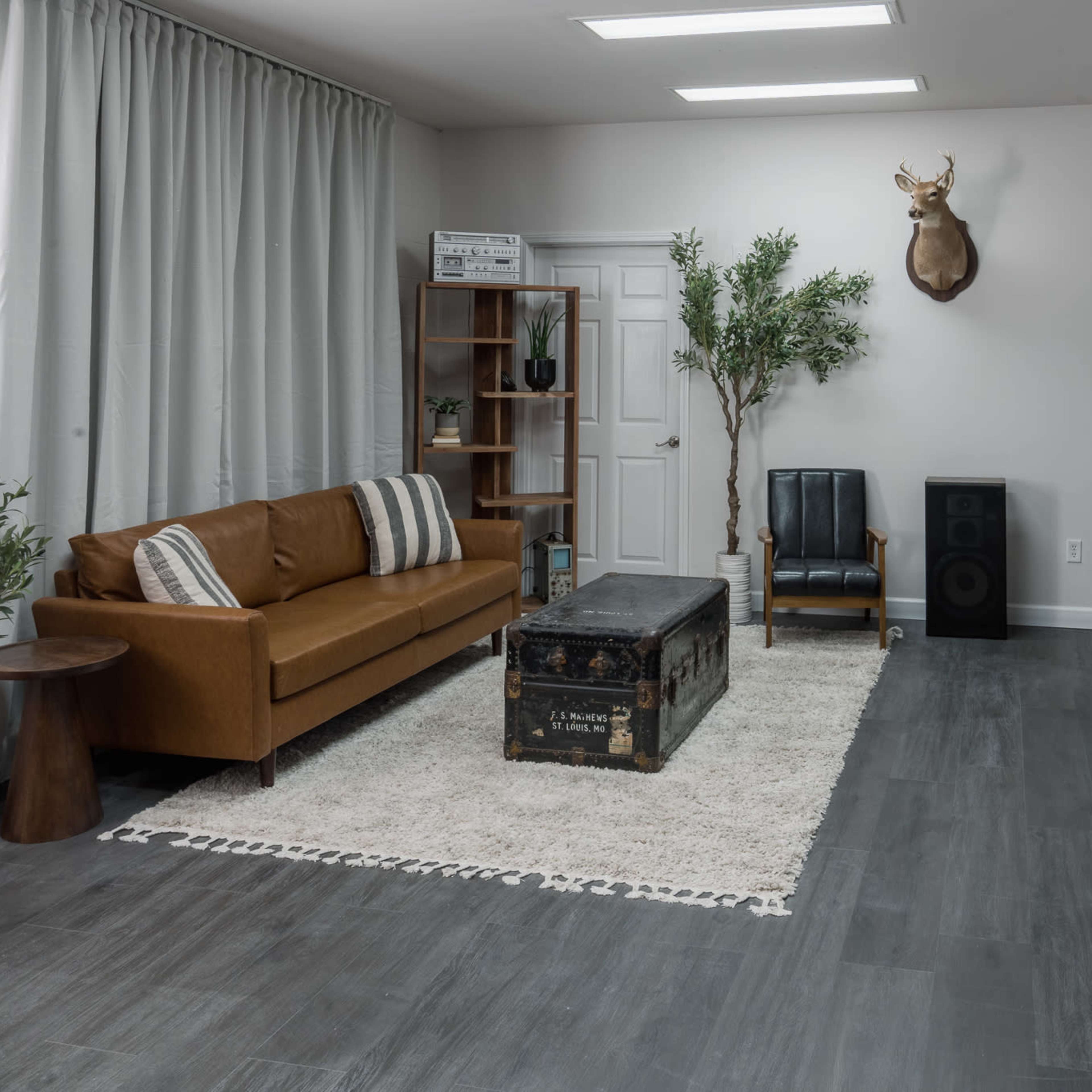 A modern living space featuring a brown leather sofa, a vintage trunk coffee table, and a deer head mounted on the wall, surrounded by greenery and a flat-screen TV.