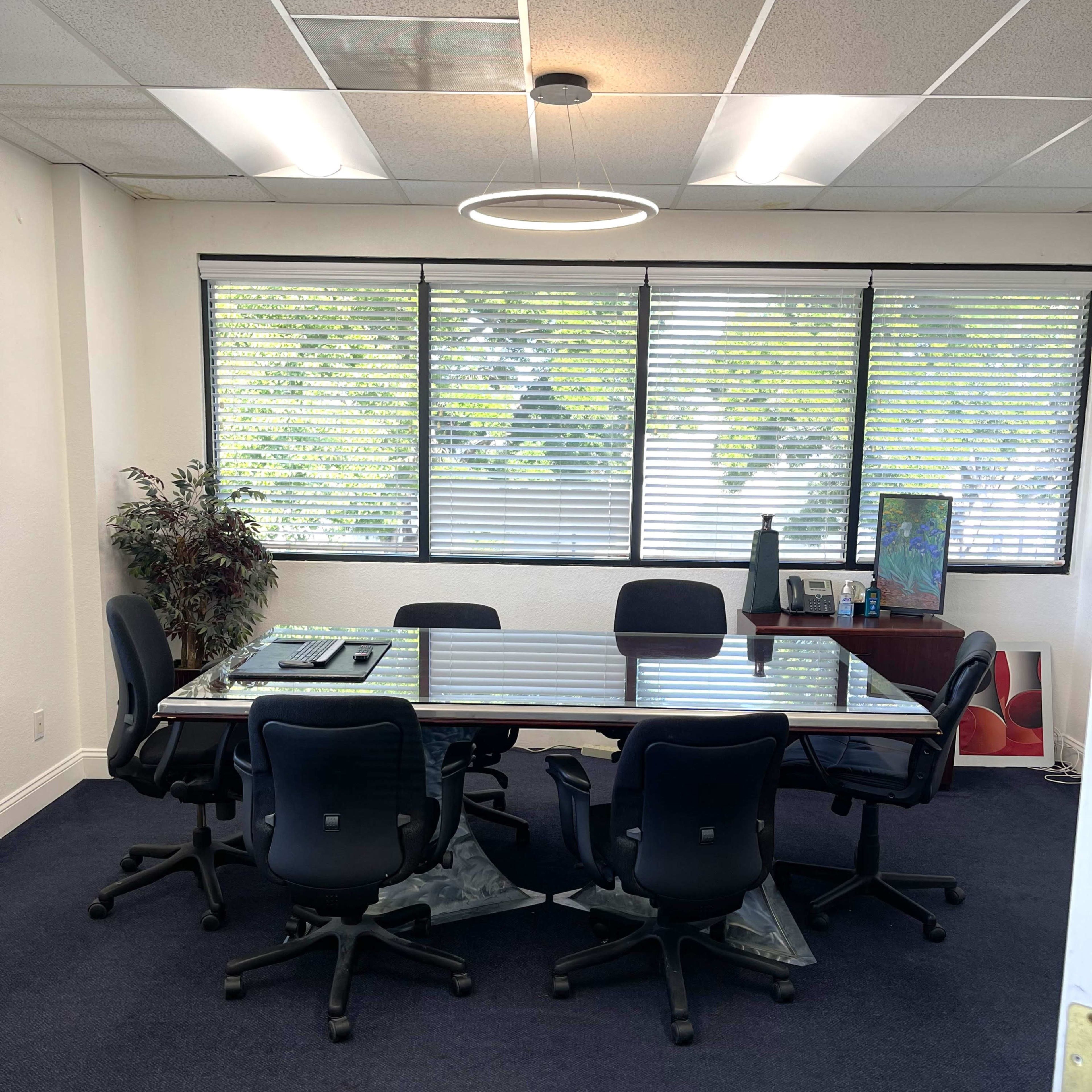 A conference room features a large glass table surrounded by six black ergonomic chairs, with large windows providing natural light.