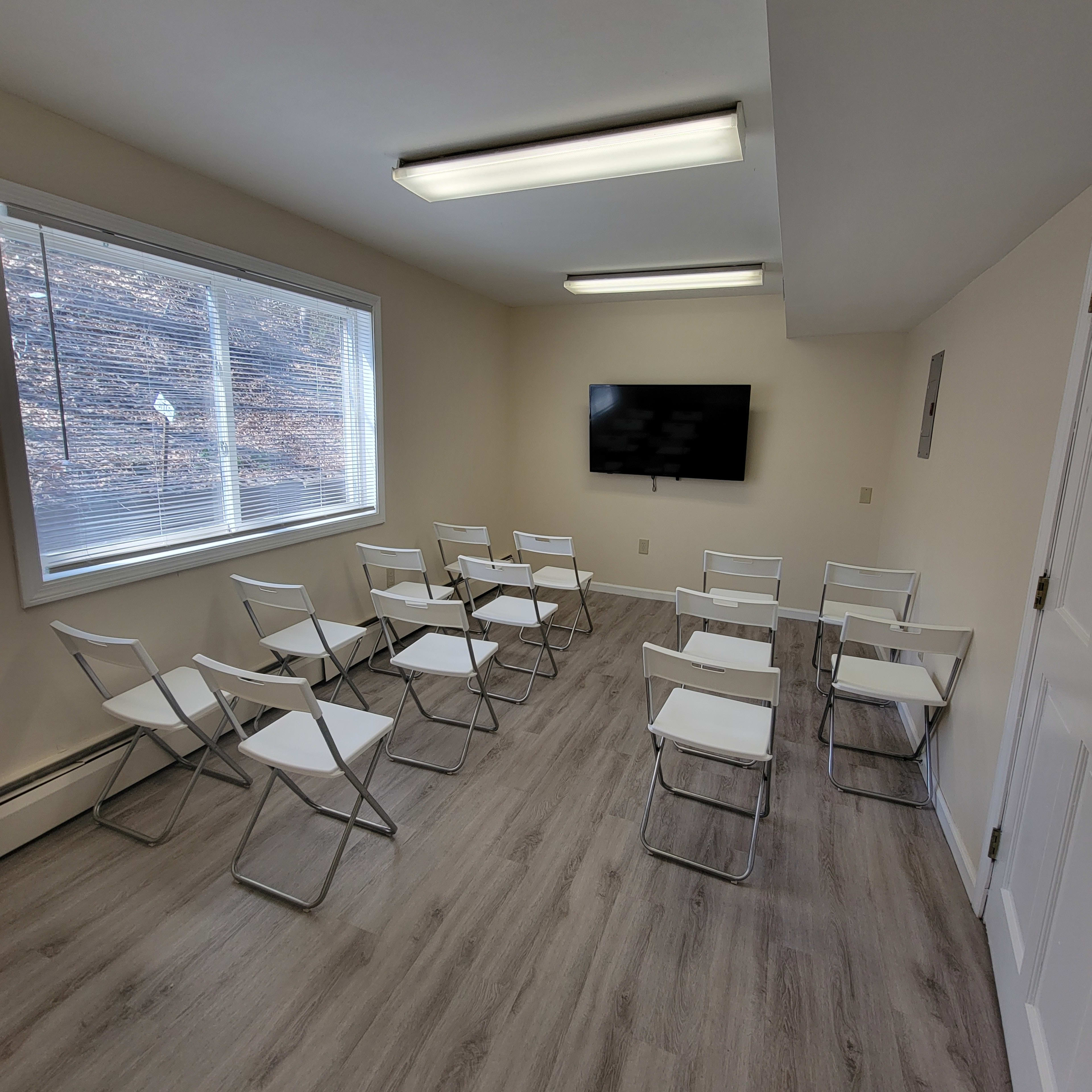 A small meeting room with white folding chairs arranged in rows facing a wall-mounted television and a large window providing natural light.